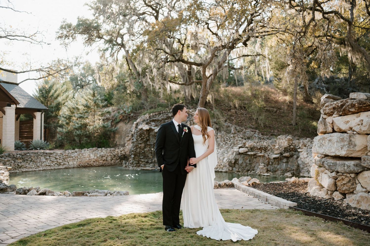 A couple stands together on stone patio surrounded by Spanish moss draped trees at an elegant outdoor venue.