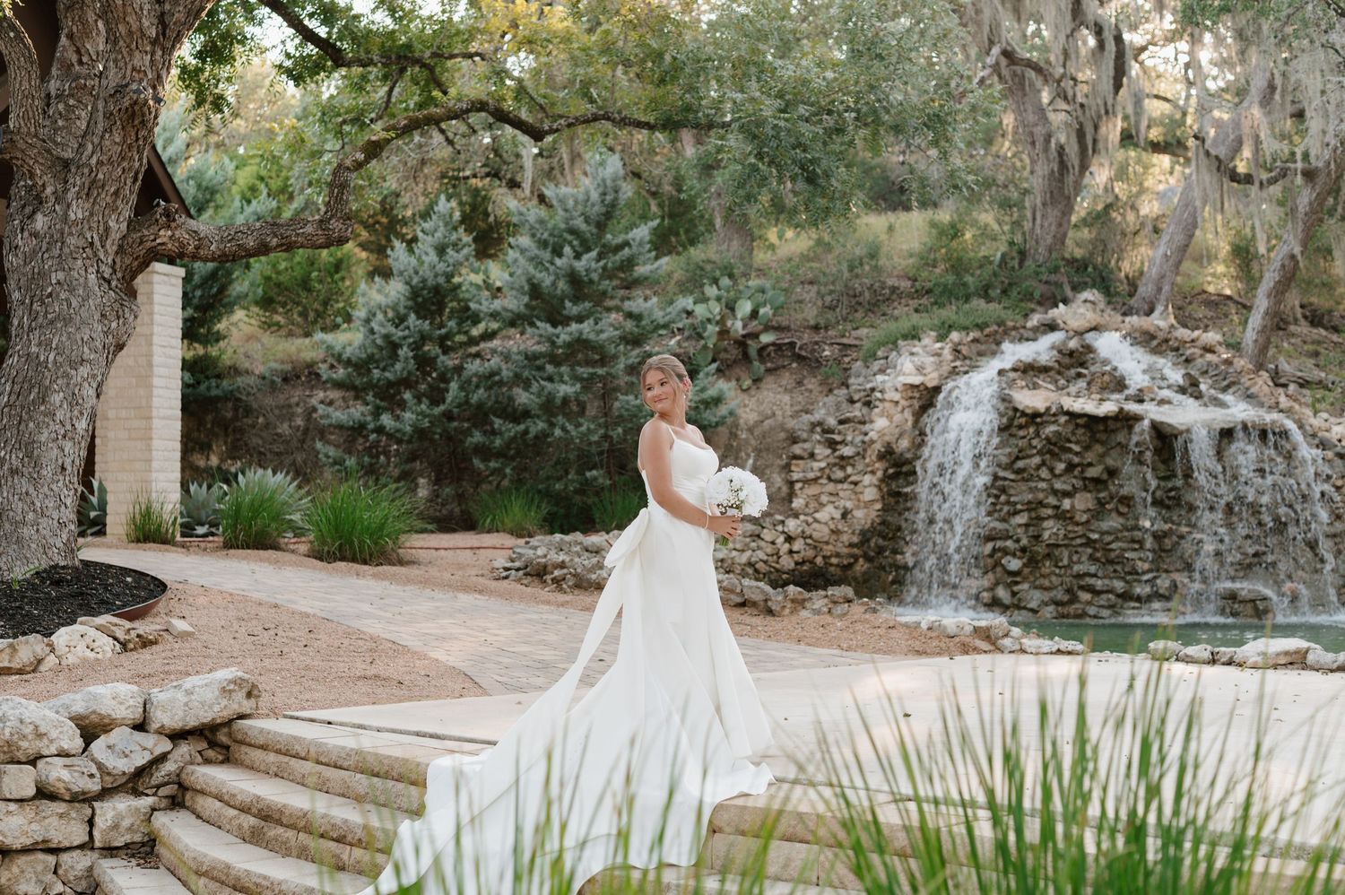 A wedding party poses in a lush garden setting with stone steps and a cascading waterfall in the background.