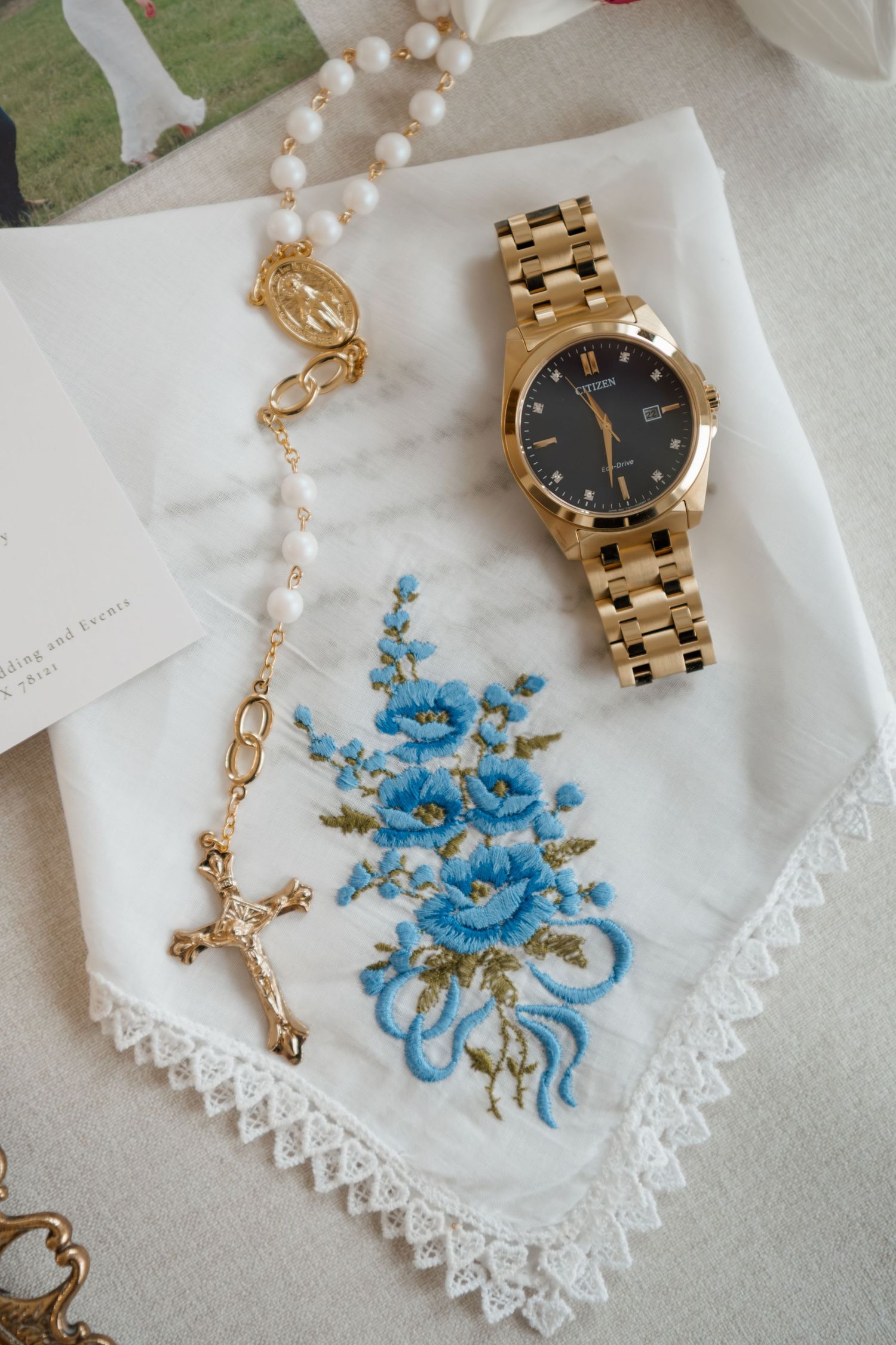 Gold watch and pearl necklace with blue floral embroidery on white lace handkerchief displayed on table.
