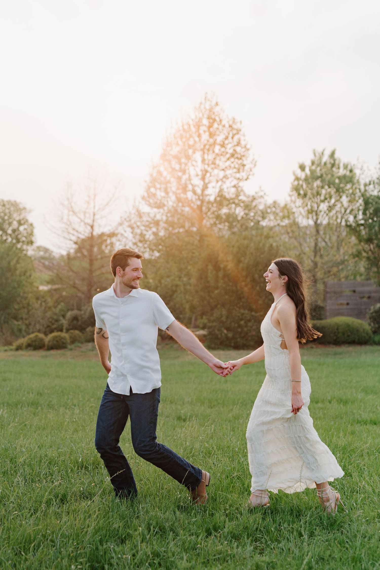 A couple holds hands while playfully walking together in a sunlit grassy meadow at sunset.