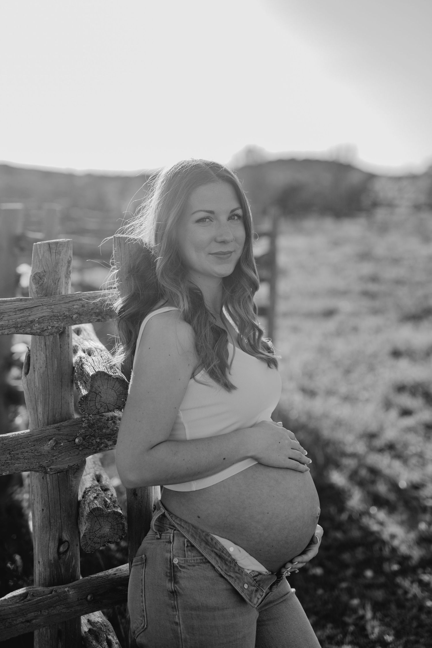 A black and white maternity portrait shows an expectant mother in a tank top leaning against a rustic wooden fence.