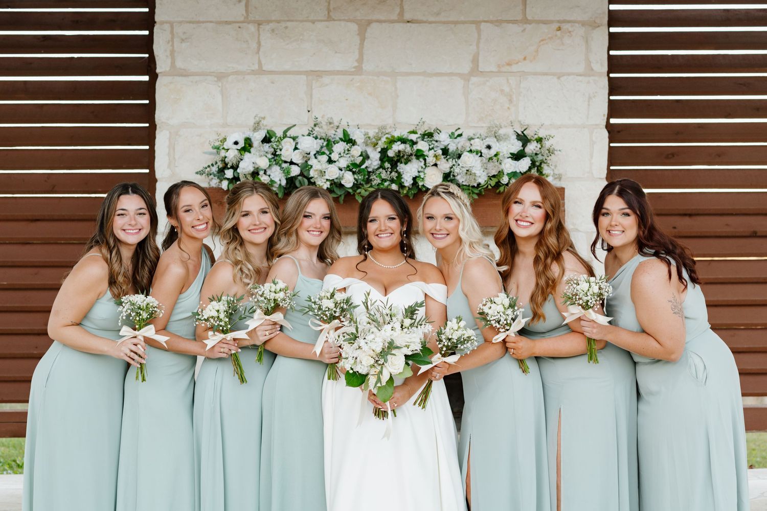Wedding party wearing seafoam green bridesmaid dresses poses together with white floral bouquets against wood wall.
