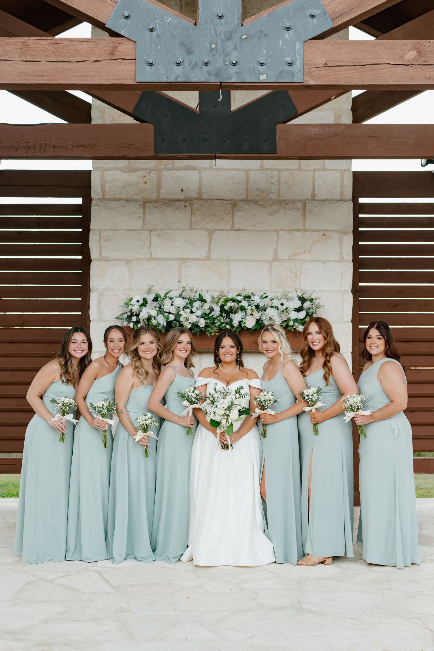 A bridal party wearing soft mint green bridesmaid dresses poses together under a rustic wooden pavilion.