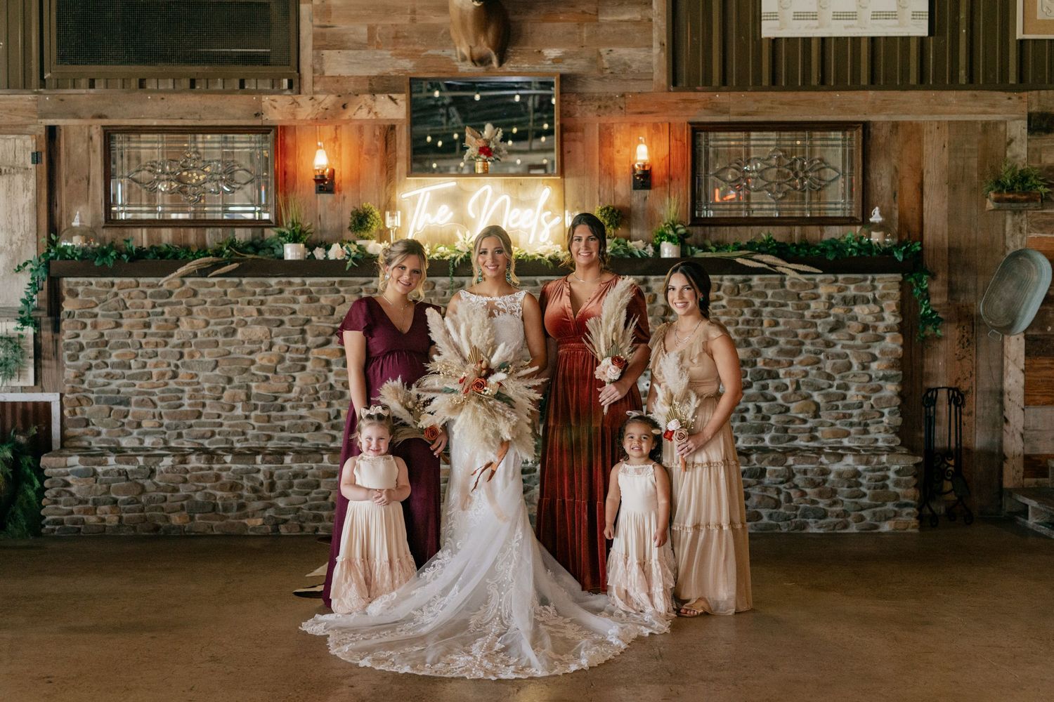 Elegant wedding party gathered in front of stone wall with warm lighting and decorative greenery.
