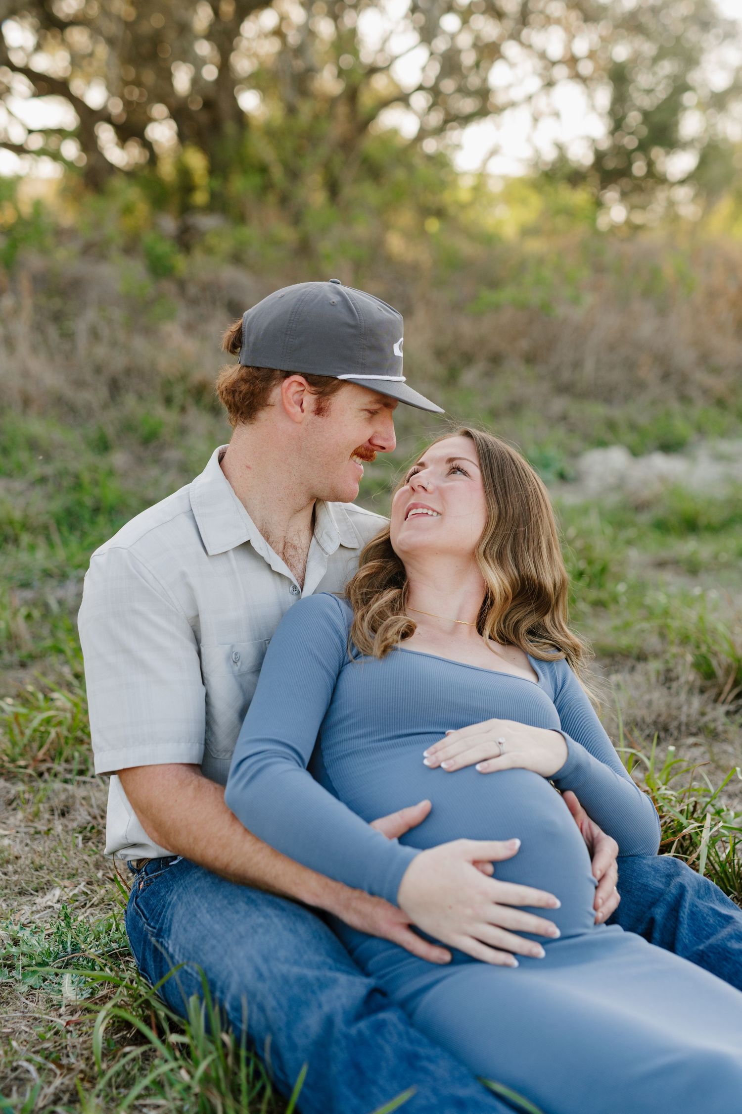 Intimate maternity photo session shows a couple sitting in grass as the sun sets behind them.