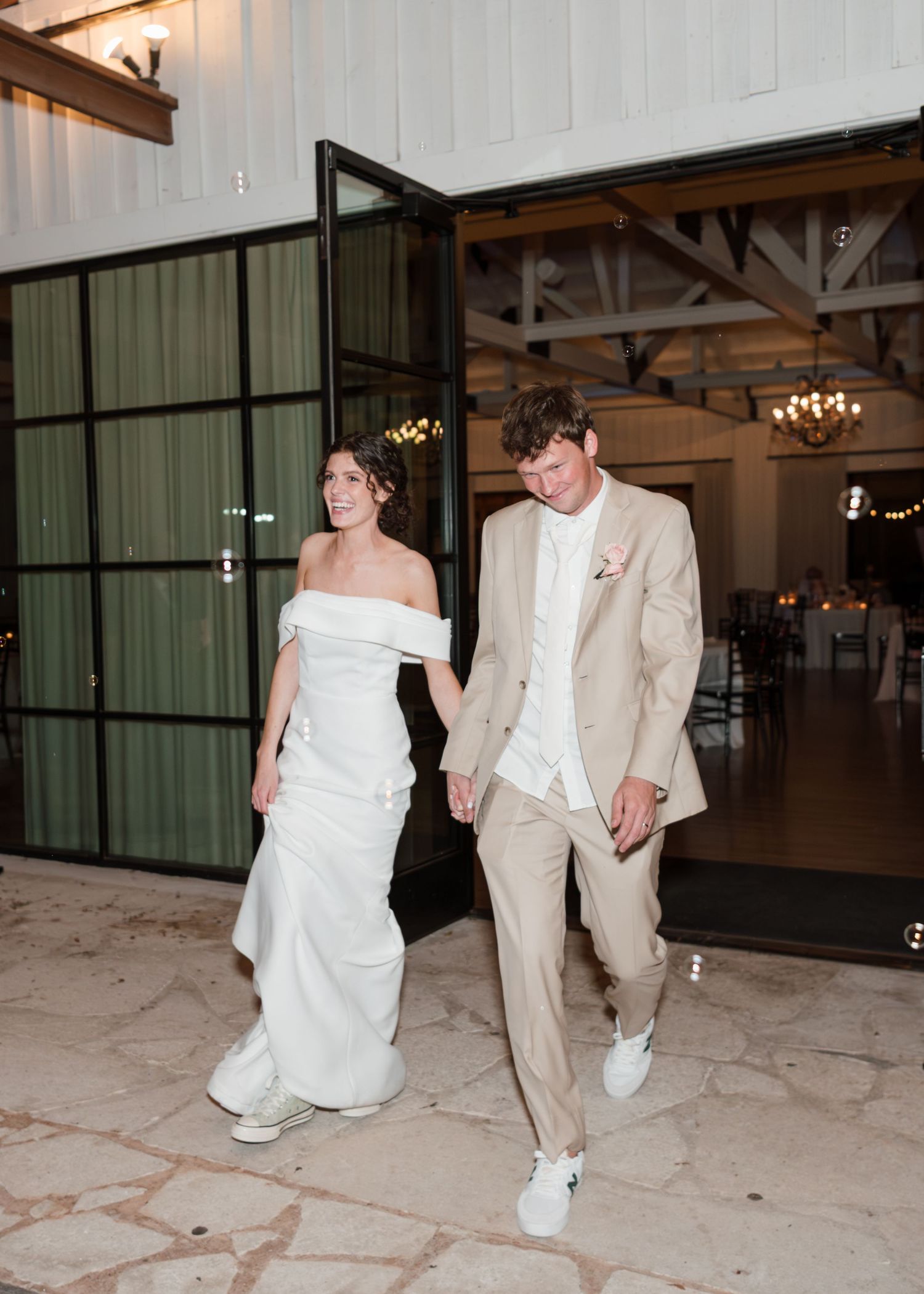 Newlywed couple exits venue holding hands, bride in white dress and groom in tan suit with sneakers.