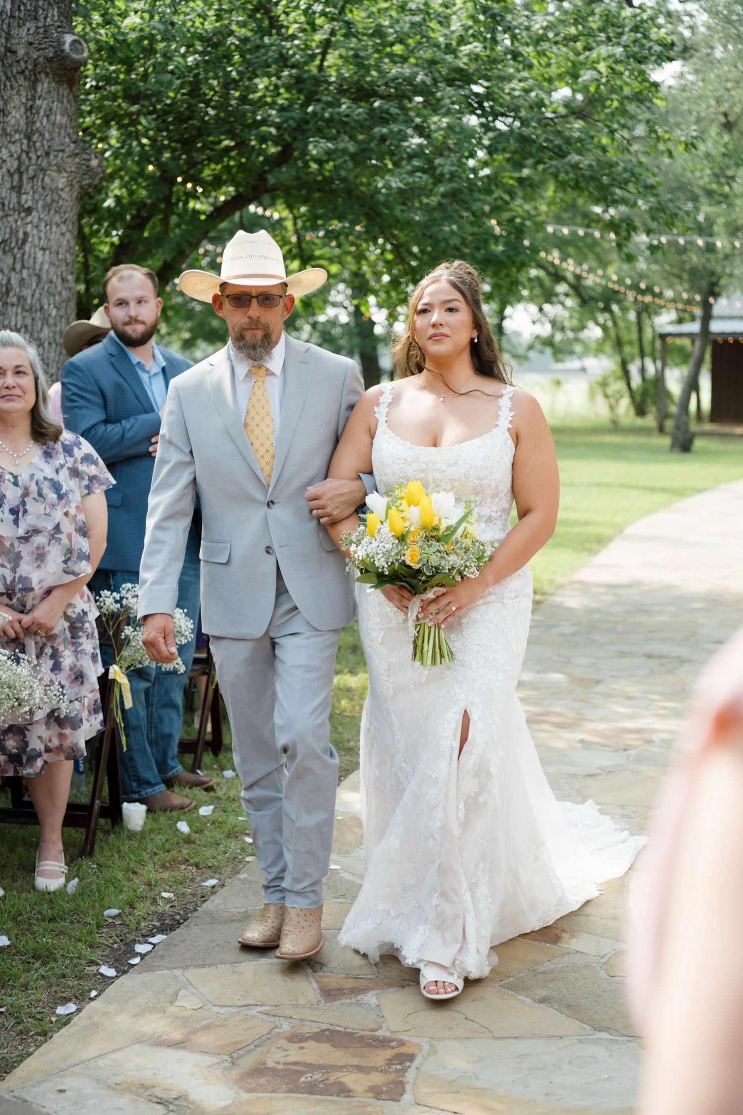 Bride in lace gown holding yellow bouquet walks down outdoor aisle with man in gray suit and cowboy hat.