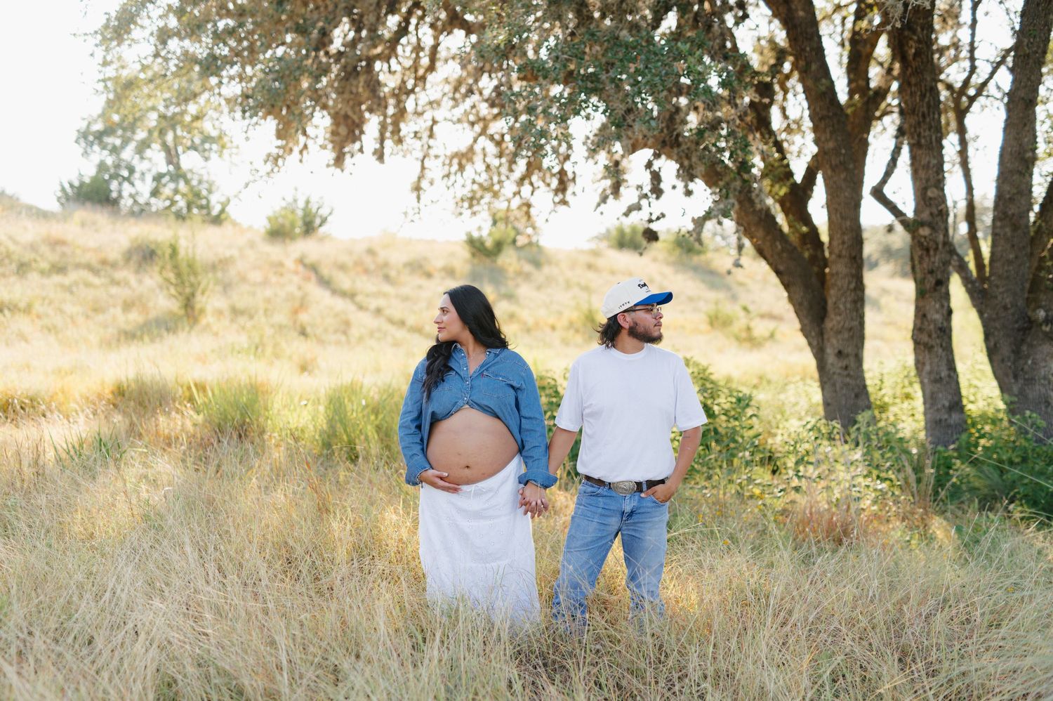 Expectant couple poses for photos along a scenic path with golden sunlight filtering through trees.