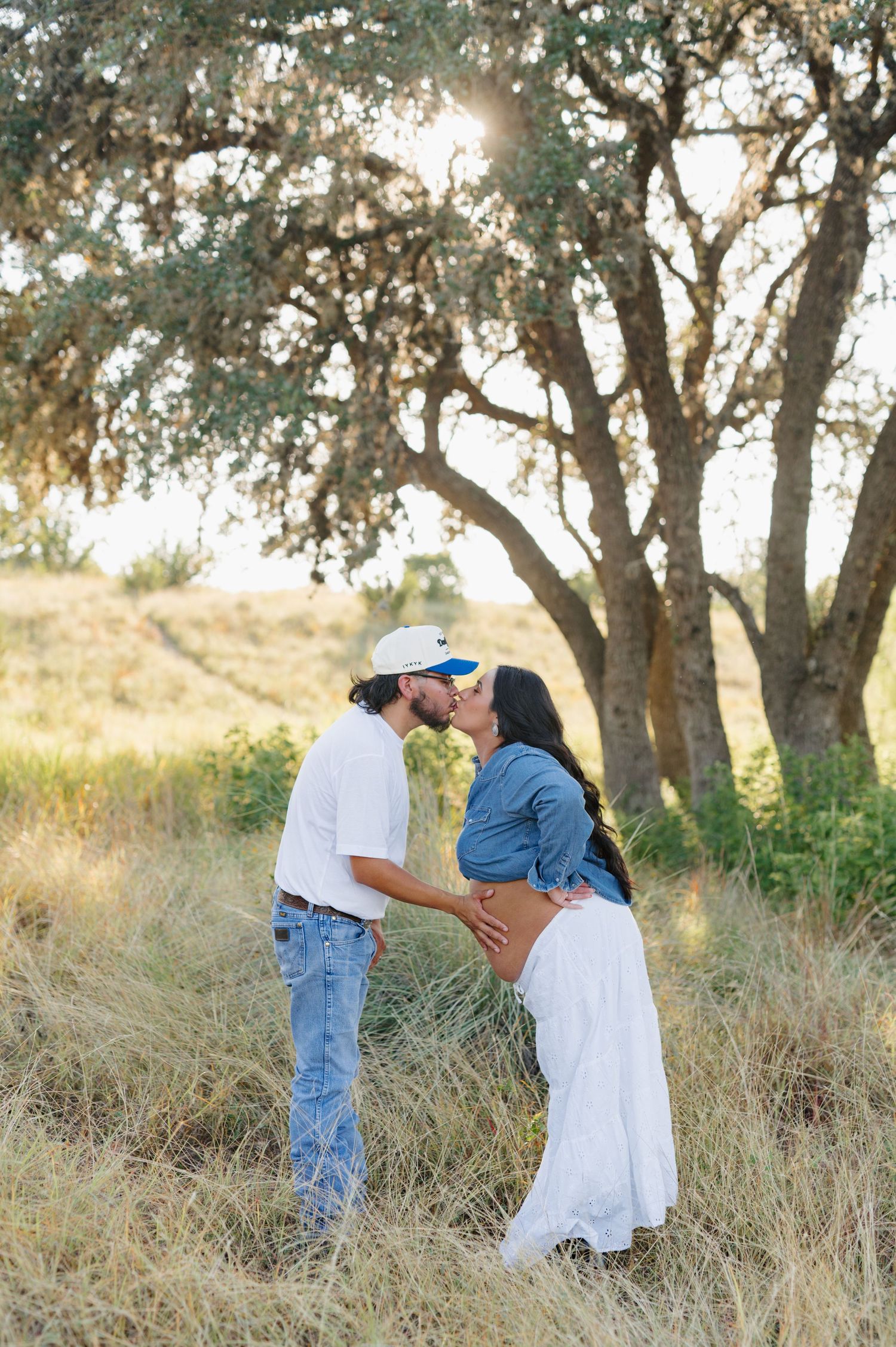 A couple shares an intimate moment among oak trees in a sunlit natural setting.