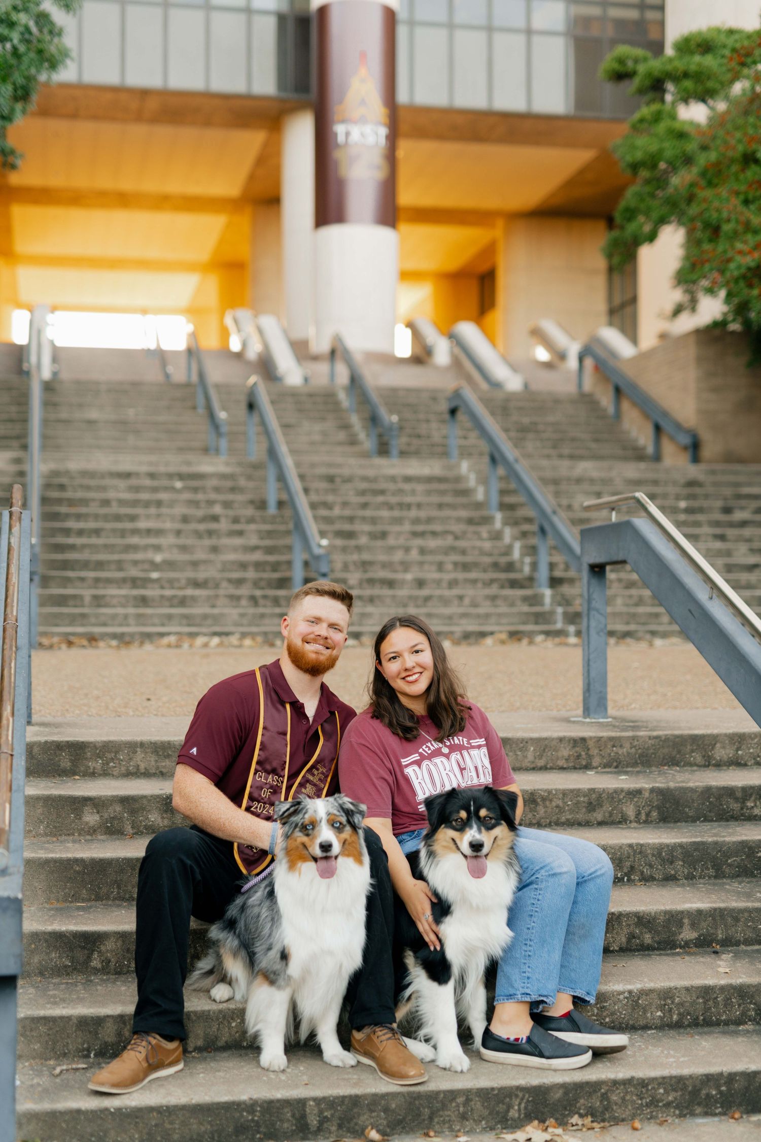 Couple sits on concrete steps with two Australian Shepherds wearing matching maroon university attire.