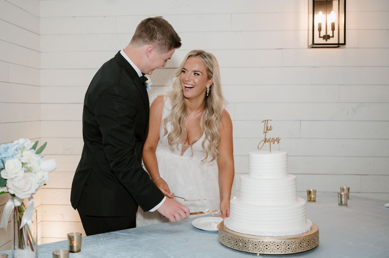 A newlywed couple stands next to a large white wedding cake, the bride in a flowing white dress and the groom in a black suit.
