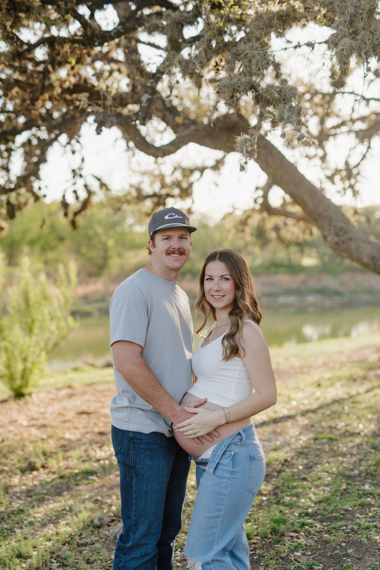 A couple stands together under a large tree branch during golden hour in a natural outdoor setting.