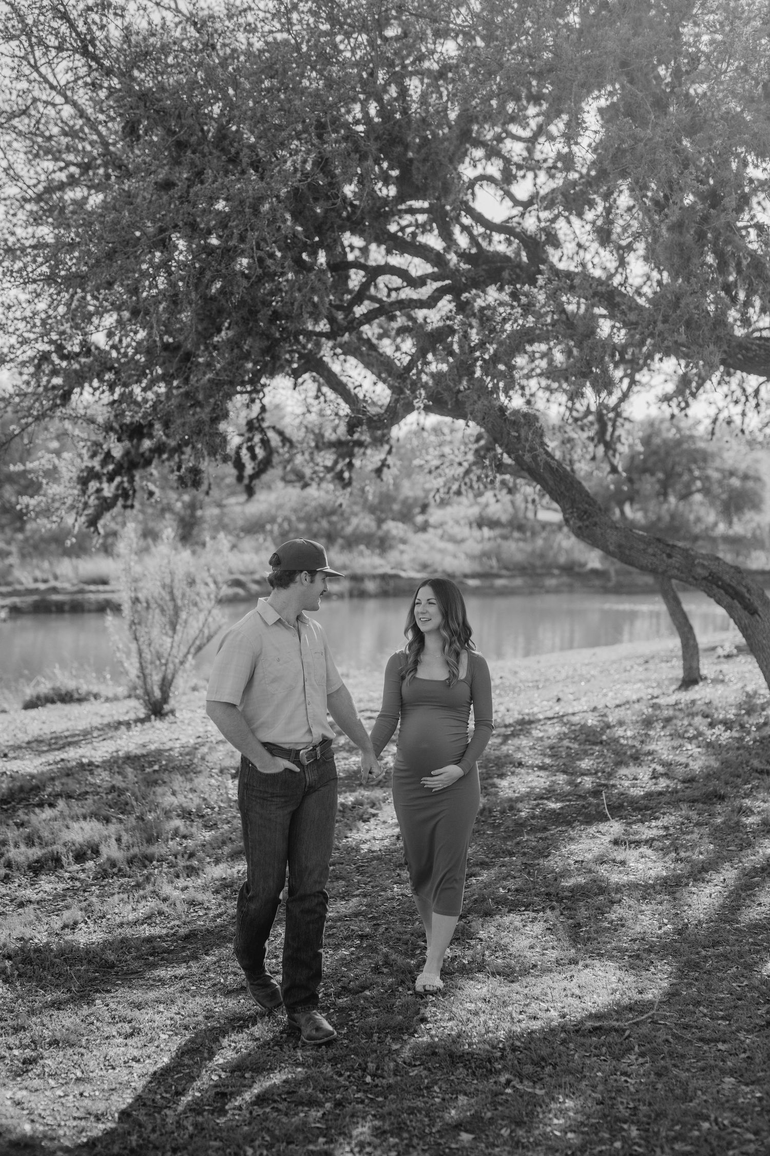 A black and white photo of an expecting couple walking together by a serene lakeside.