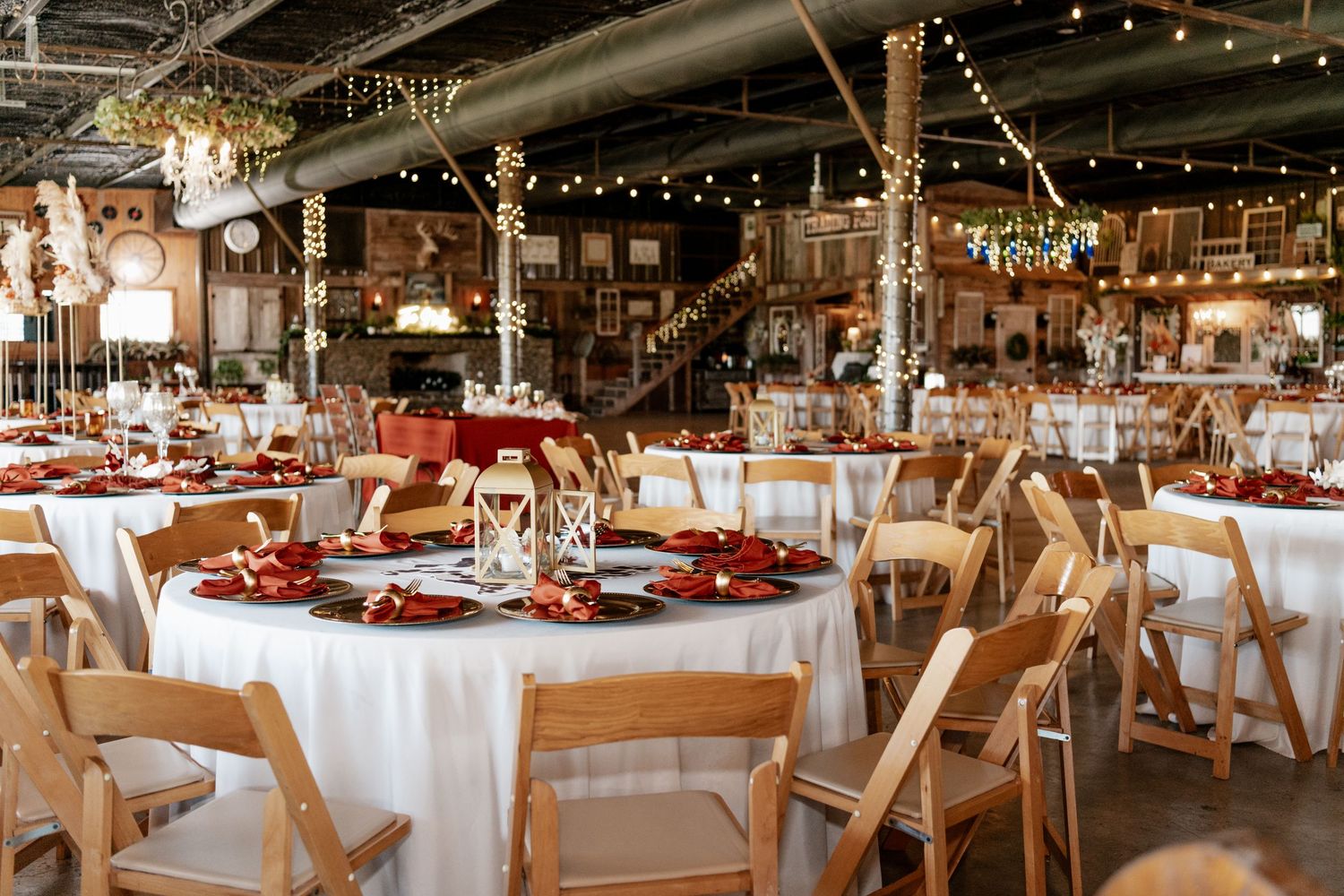Rustic barn wedding reception setup with round tables, white linens, wooden chairs and romantic string lighting overhead.