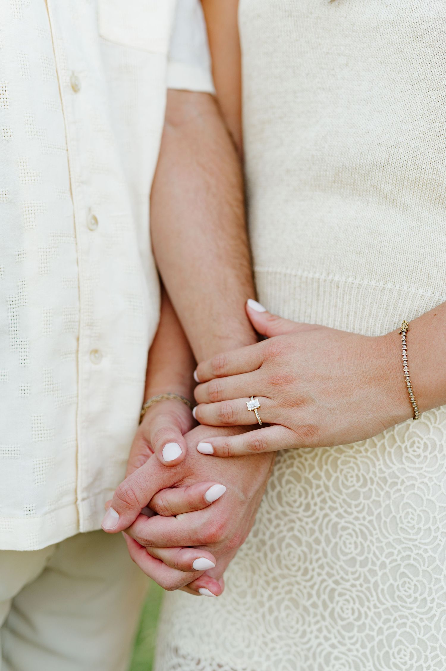 Close-up of hands holding together showing engagement rings against white clothing.