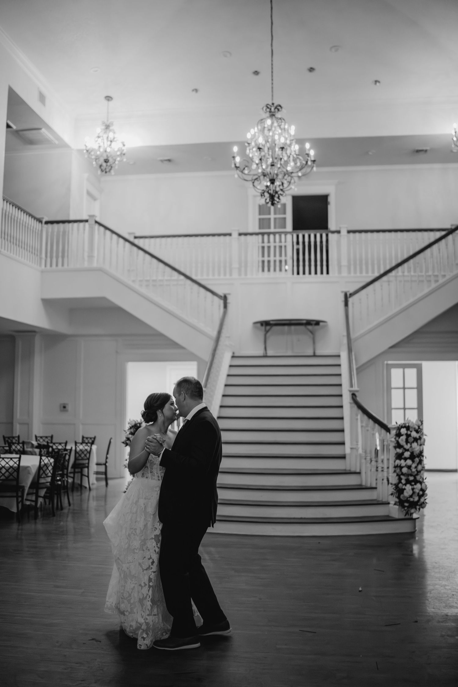 Couple dancing in grand ballroom with crystal chandeliers and symmetrical staircase.