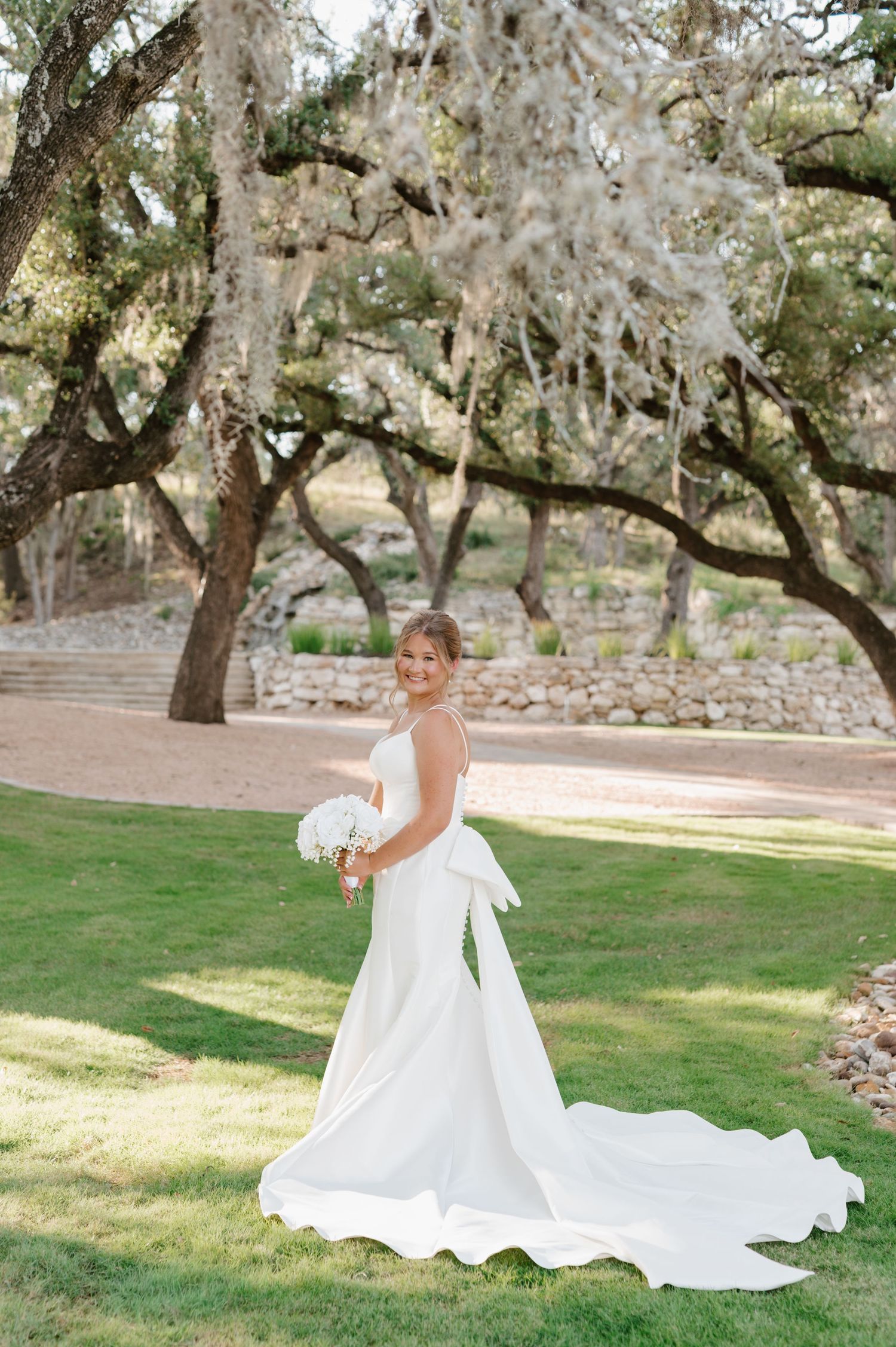 A bride in a flowing white gown poses gracefully under oak trees in a garden setting.