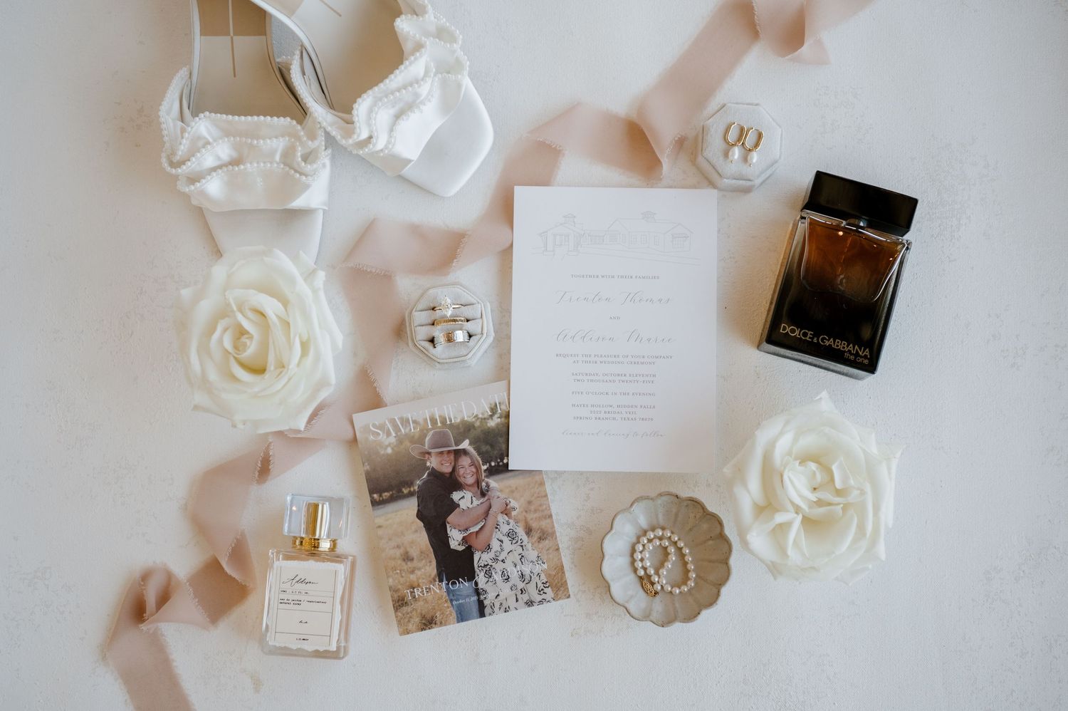Elegant wedding details including white roses, perfume bottles, and invitation cards arranged on a light surface.