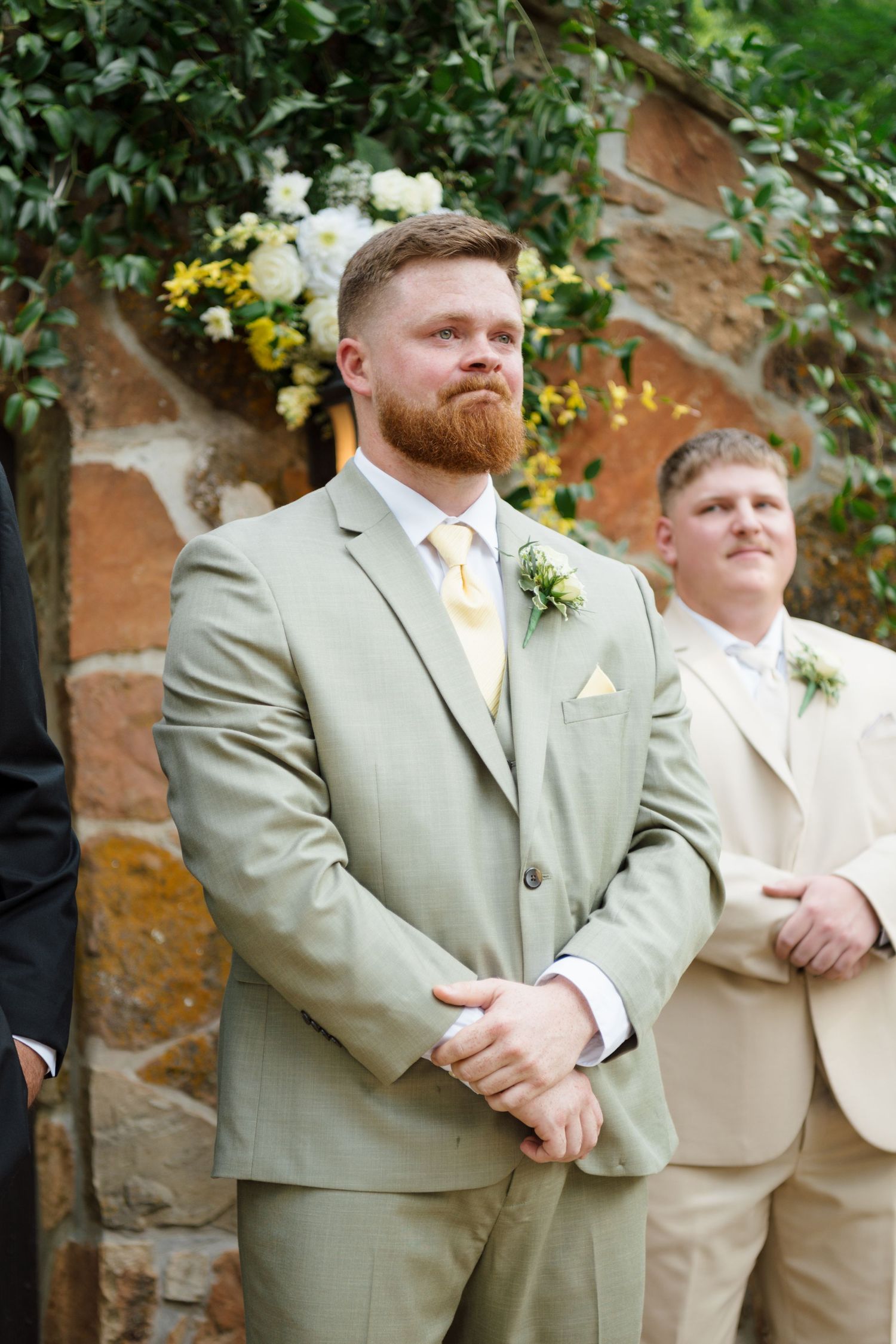 Emotional groom with red beard in sage green suit watches bride walk down aisle at outdoor wedding ceremony.