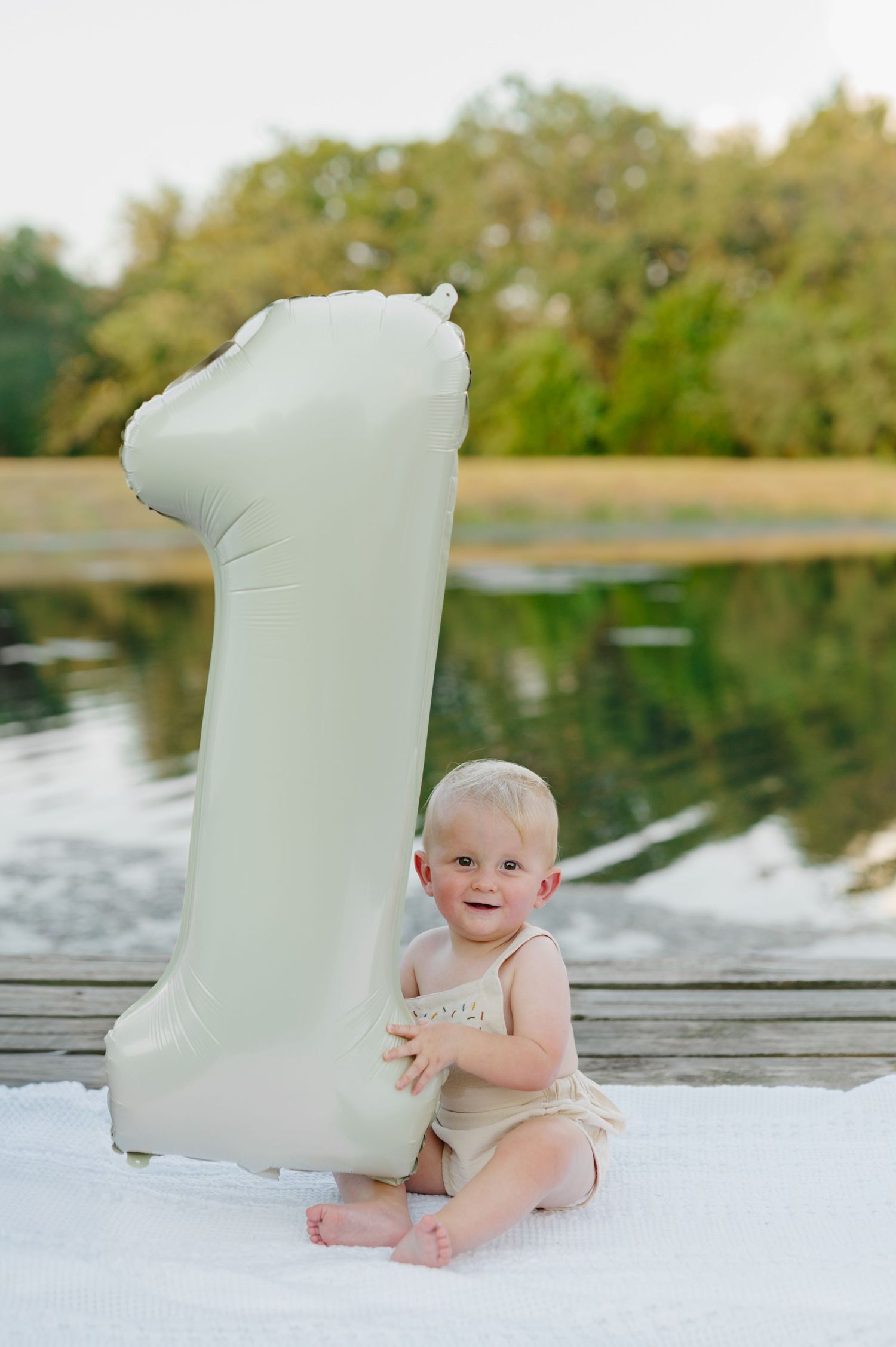 Baby sitting on wooden dock by lake with white scroll paper prop in outdoor photo session.