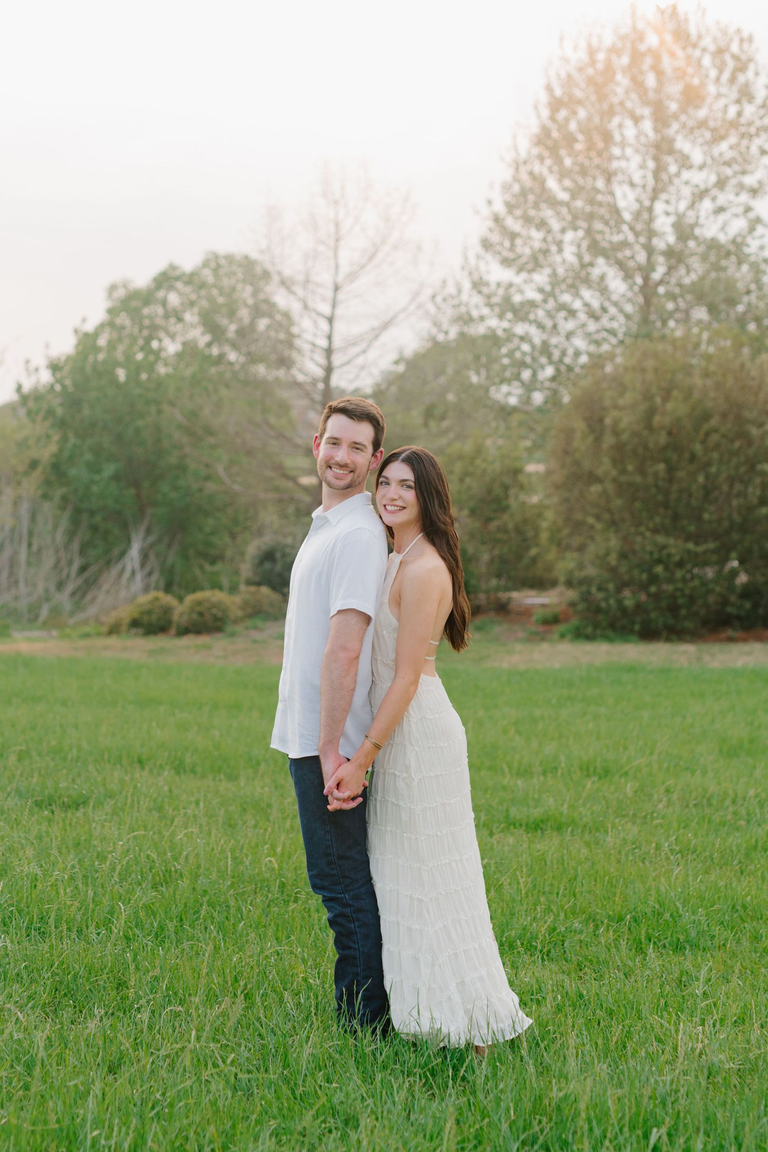 A couple poses together in a grassy field during golden hour with trees and nature in the background.