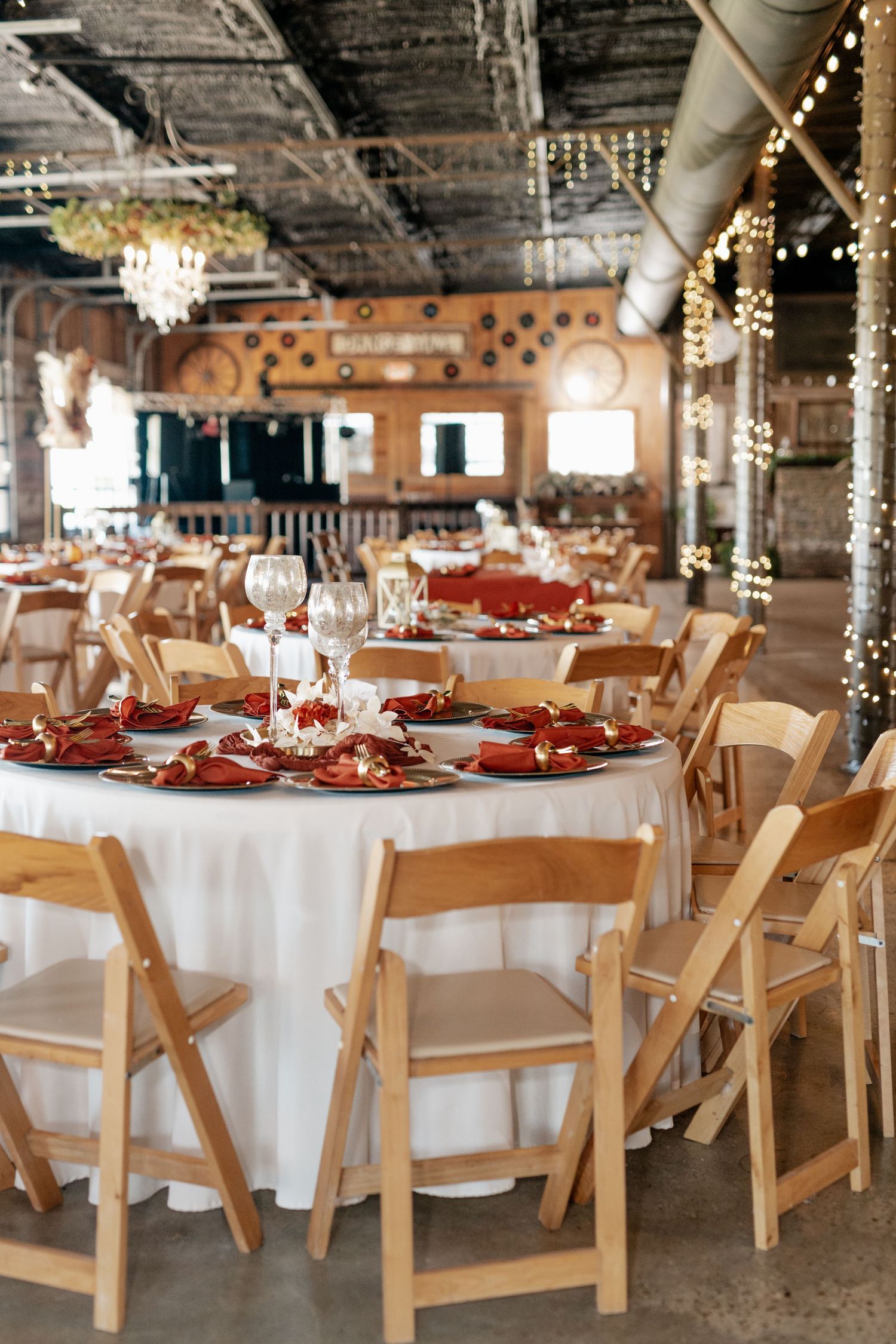 Elegant wedding reception venue with round tables set with red and white decor under string lights and chandeliers.