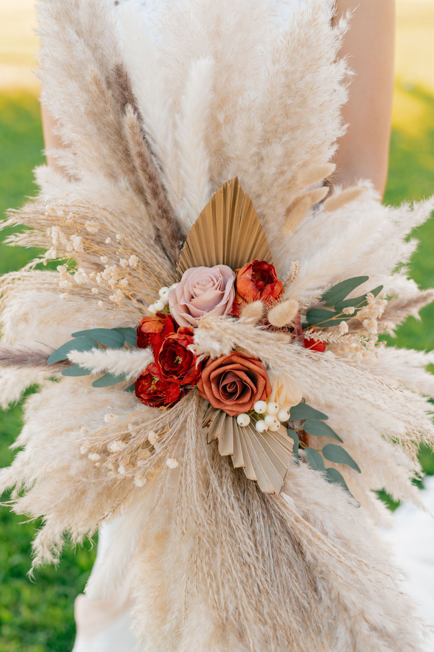 Boho wedding bouquet with dried pampas grass, pink and red roses, and palm fronds arranged in a fan shape on a sunny day.