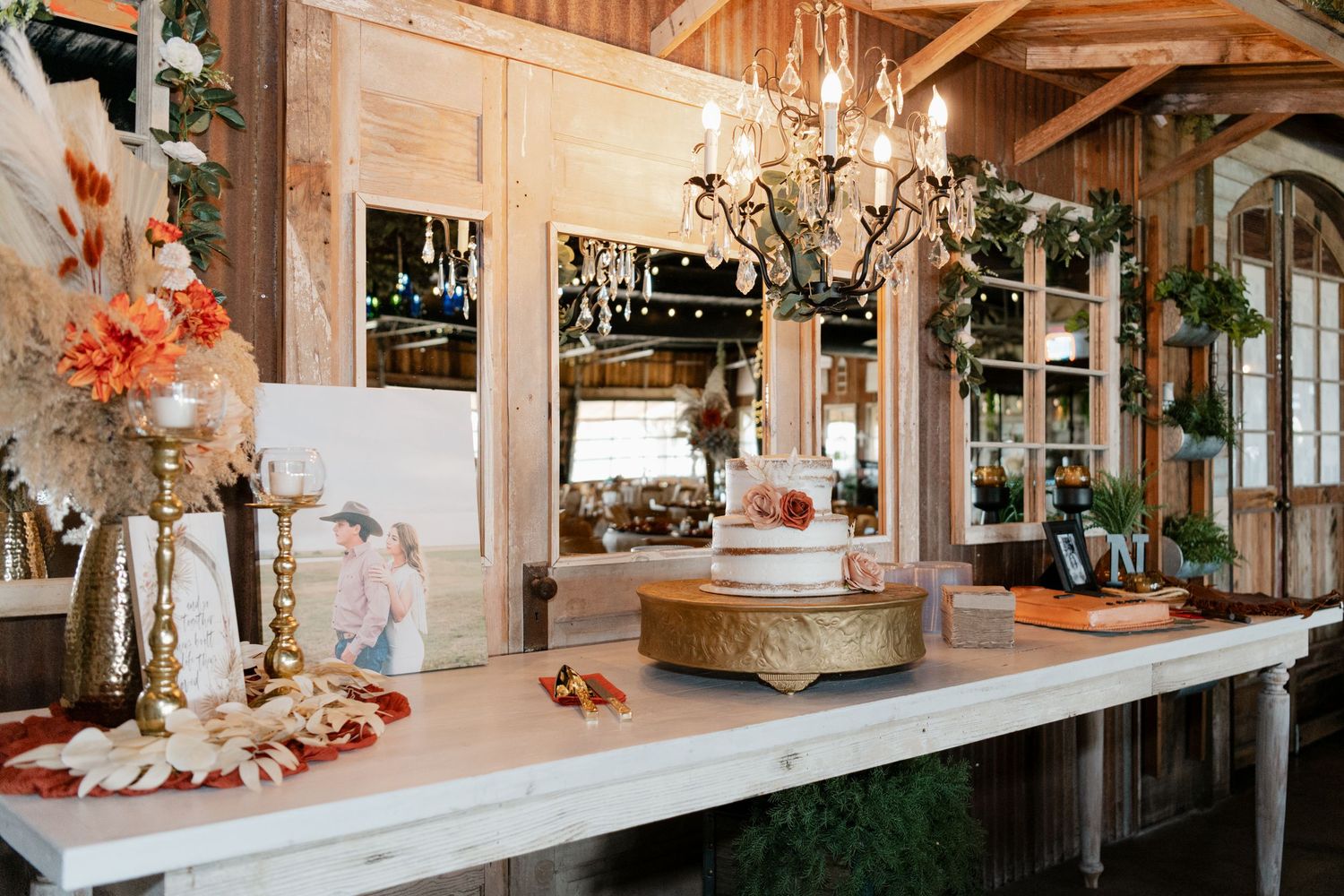Rustic wedding venue bar setup with chandeliers, mirrors, and floral arrangements on wooden counter.