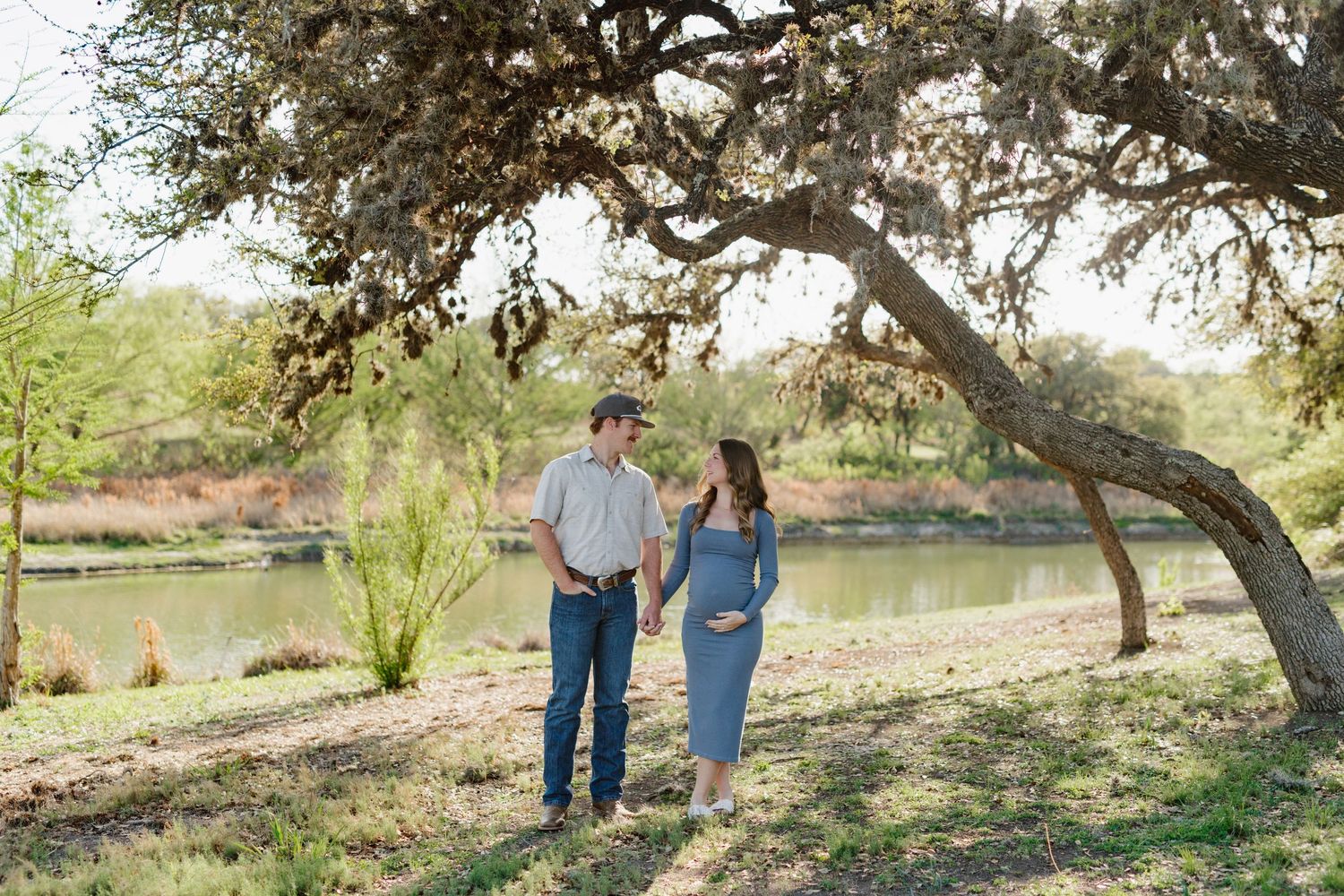 A couple strolls together under a large oak tree beside a tranquil pond on a sunny afternoon.