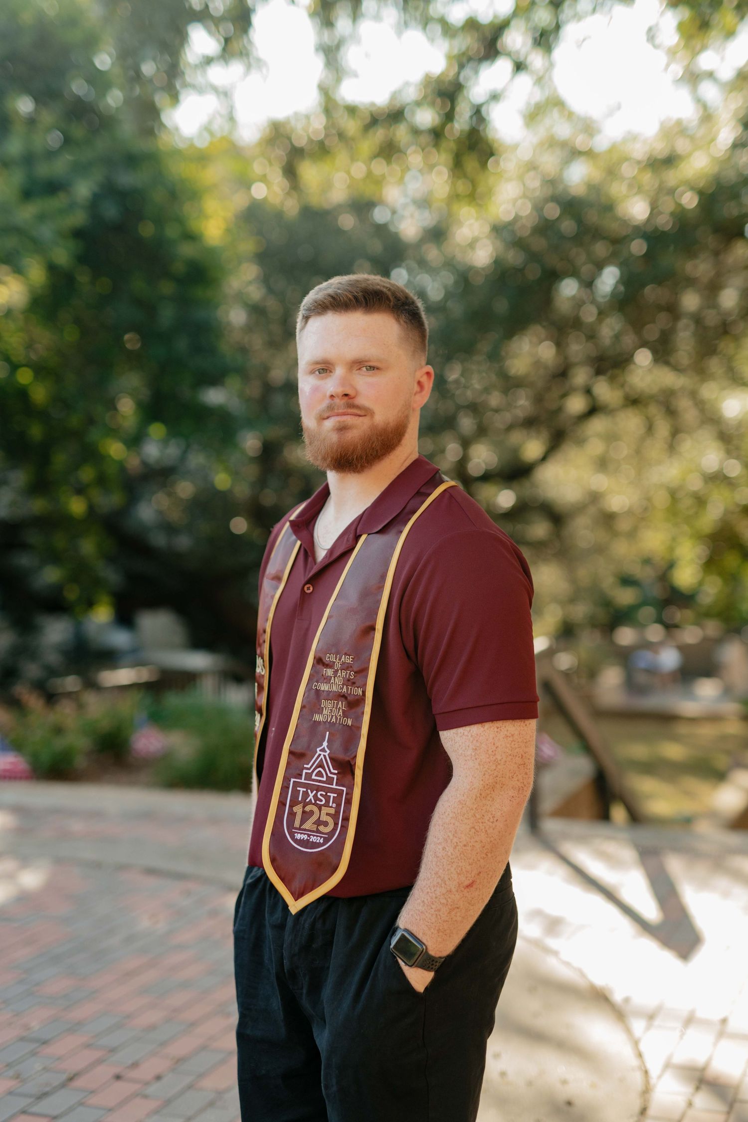 Graduate student wearing maroon academic stole and burgundy shirt poses outdoors on campus.