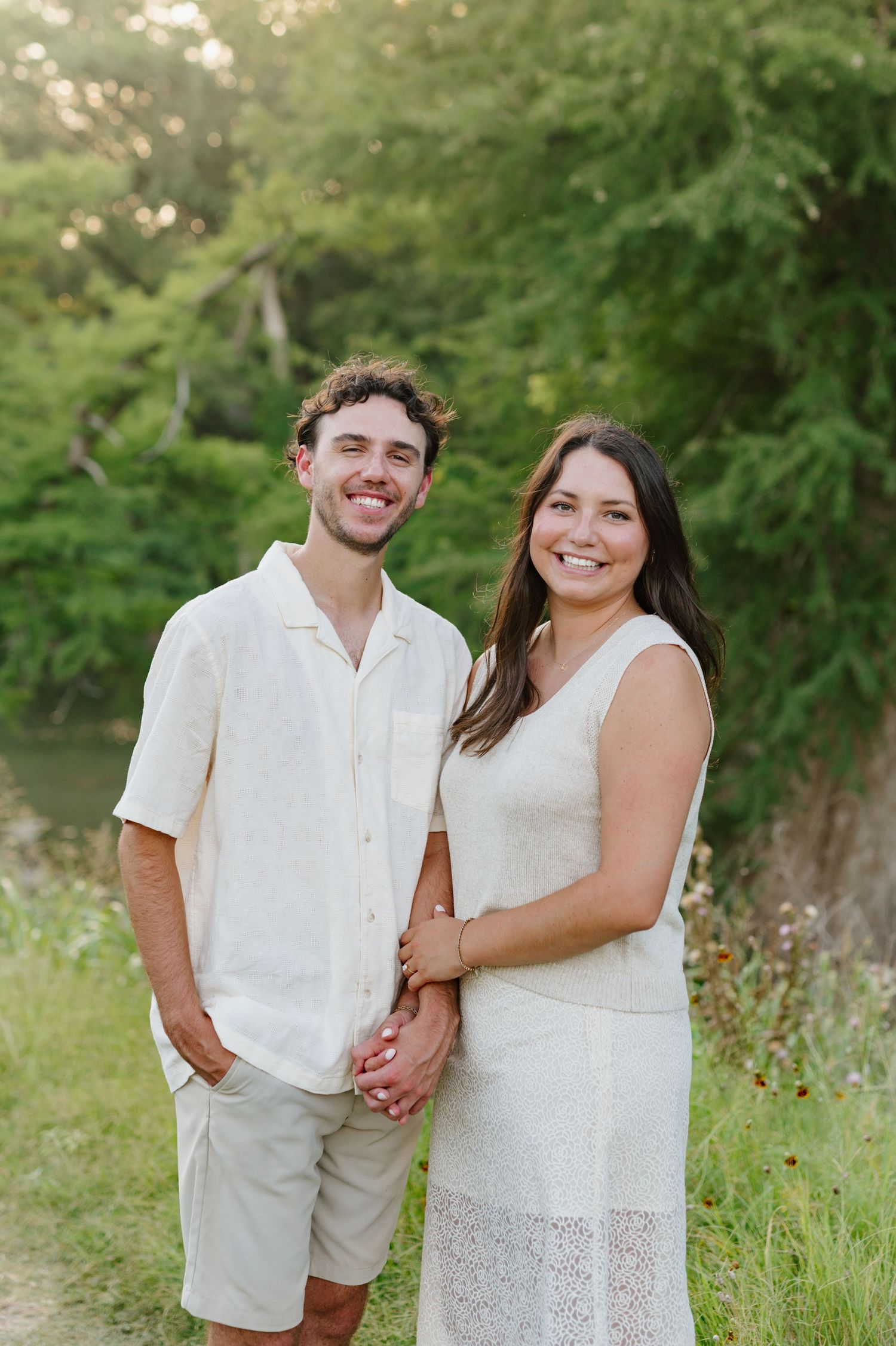 A couple in white outfits poses together in a sunny field with green foliage in the background.