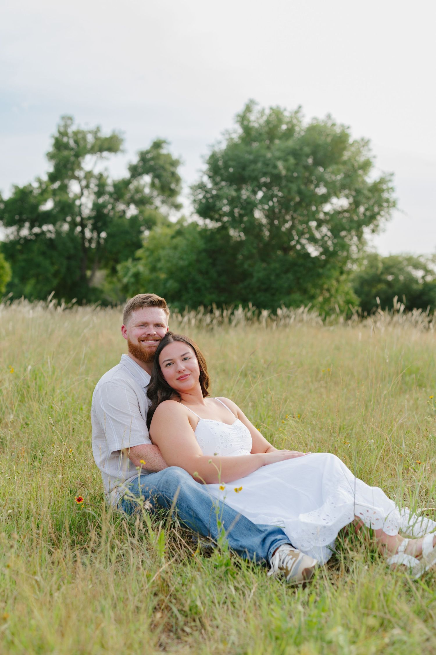 A couple rests together in a sunny field with tall grass and trees in the background.