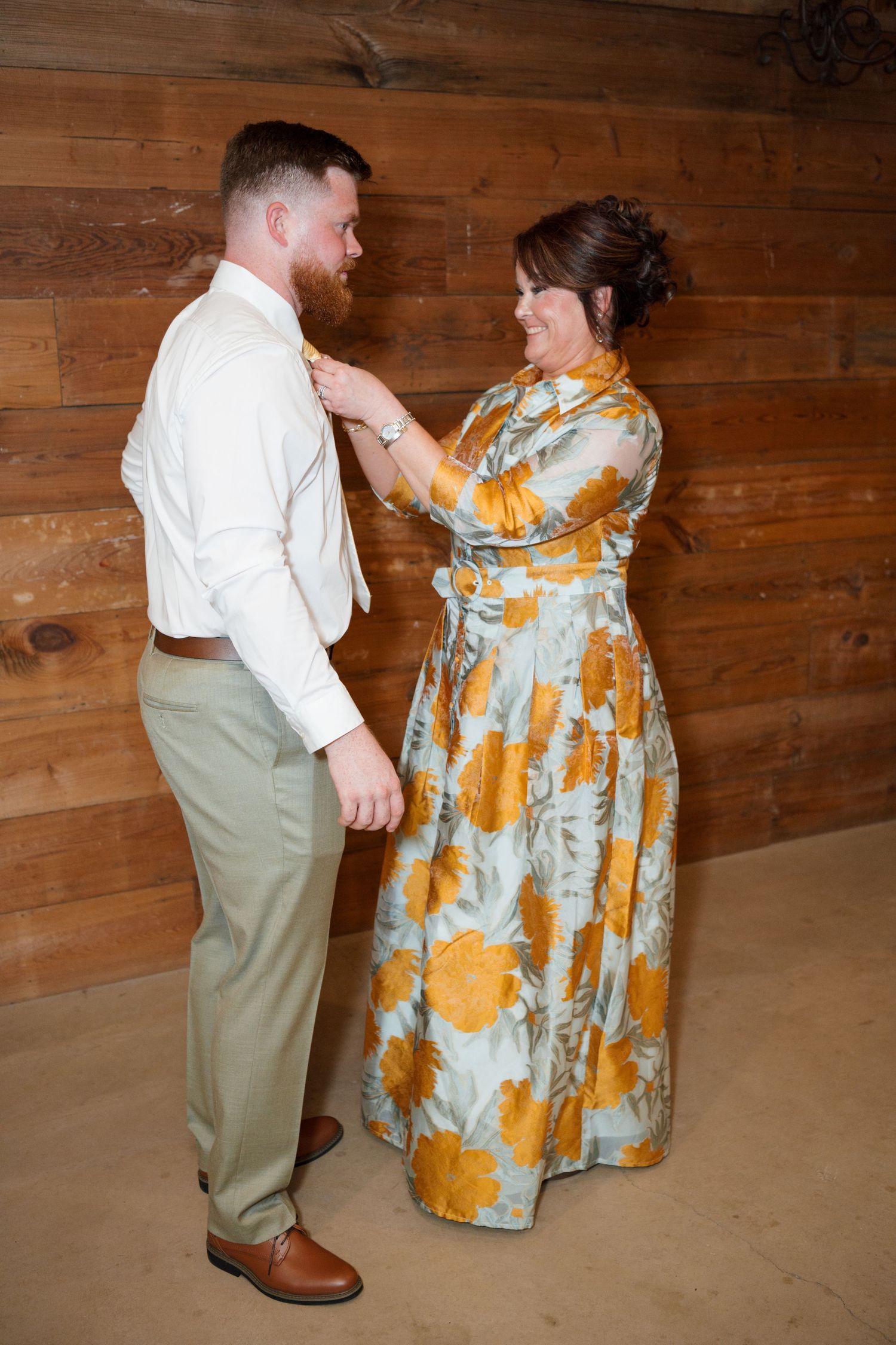 A woman in a floral gown adjusts a grooms boutonniere in a rustic wooden barn setting.