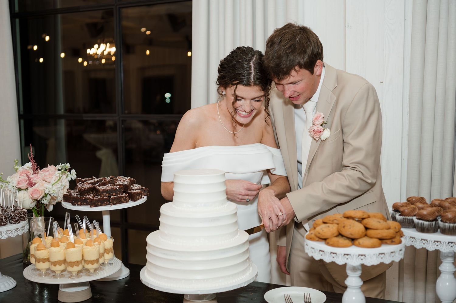 Couple cuts their classic white tiered wedding cake at dessert table during reception.