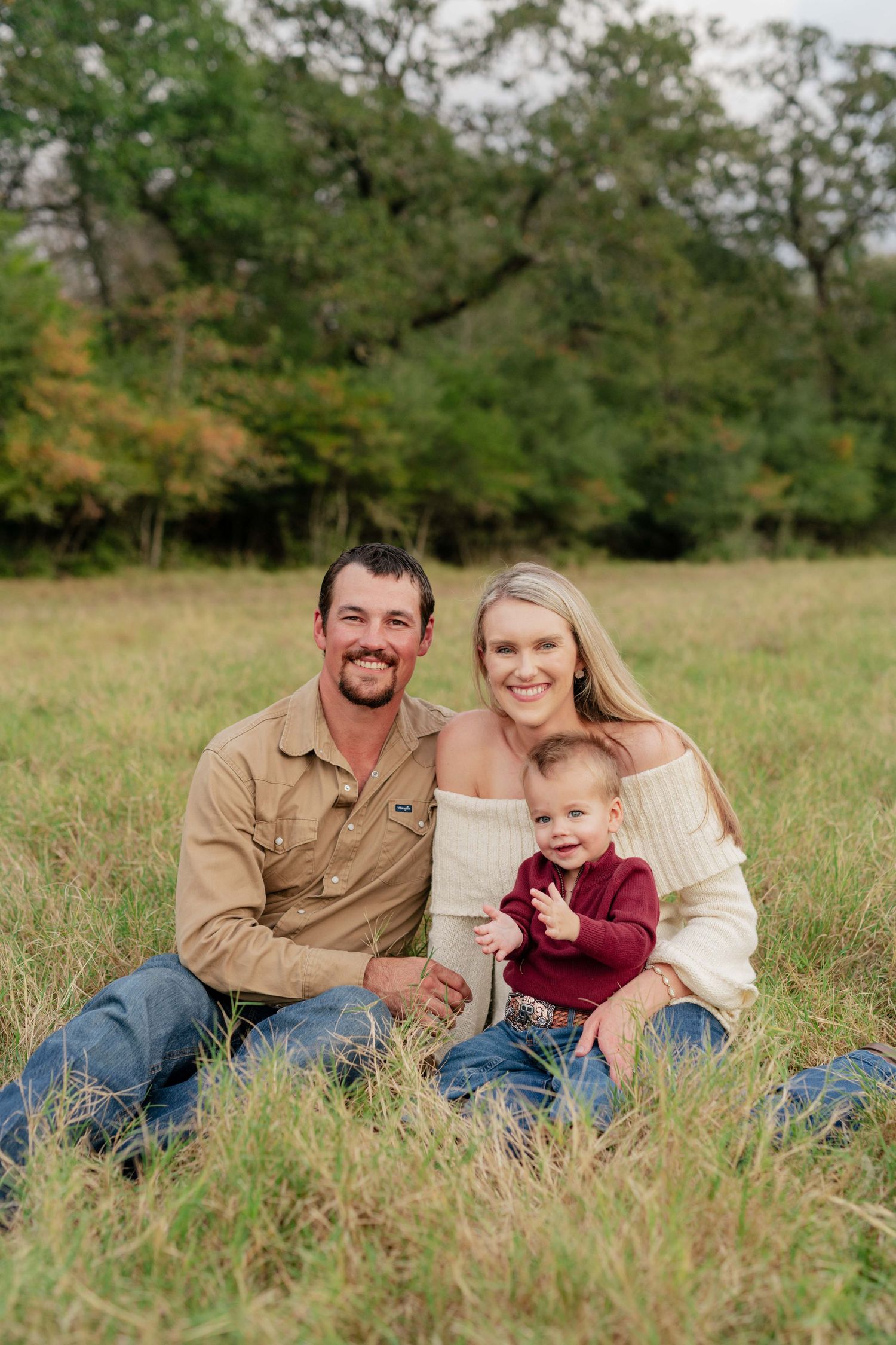 Family of three sits together on grass with trees in background during autumn photo session.