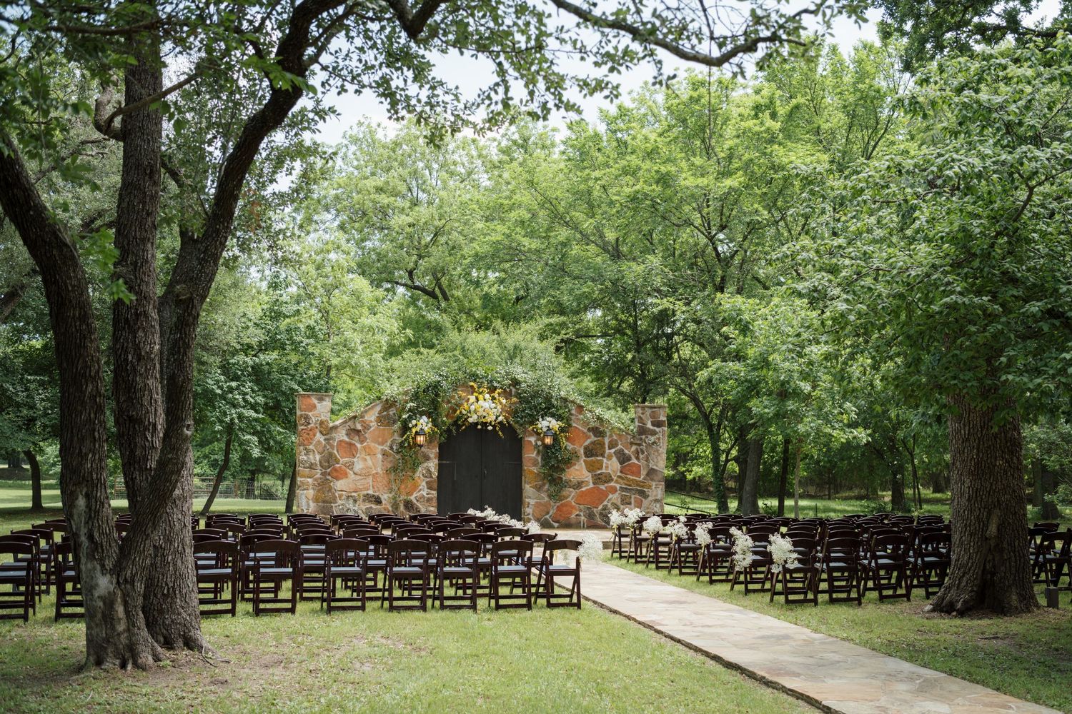 Outdoor wedding ceremony setup with dark wood chairs on a grass aisle leading to a rustic brick altar surrounded by lush trees.