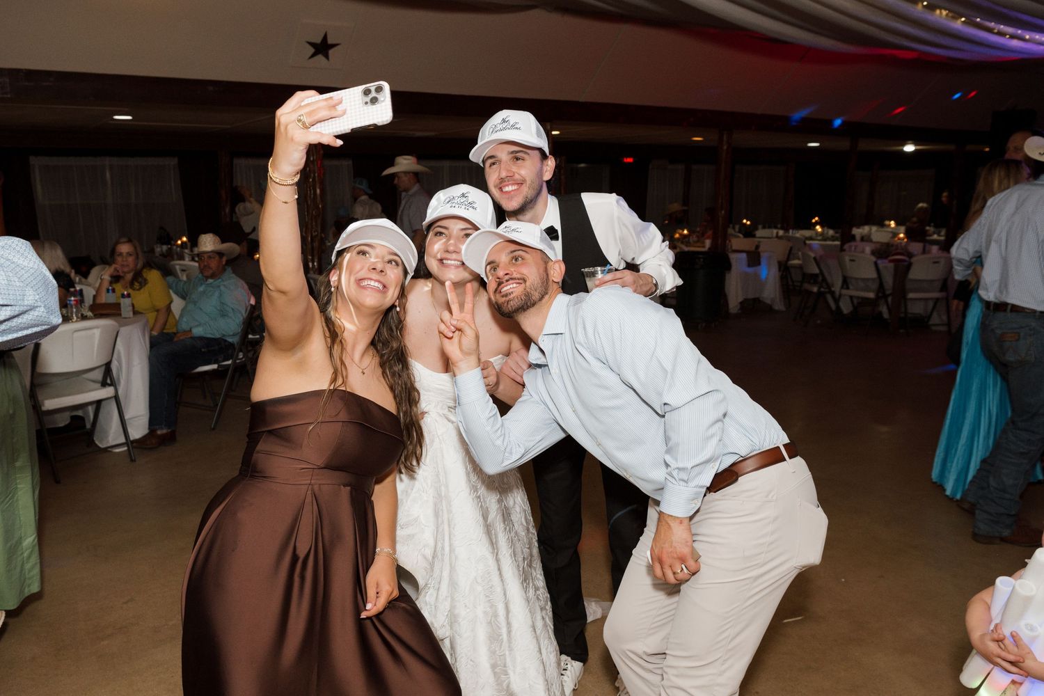 Wedding guests take fun selfie wearing matching caps at lively indoor wedding reception.