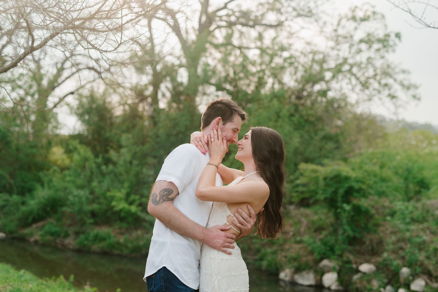 A couple embraces in a romantic kiss along a riverside path surrounded by spring greenery and trees.