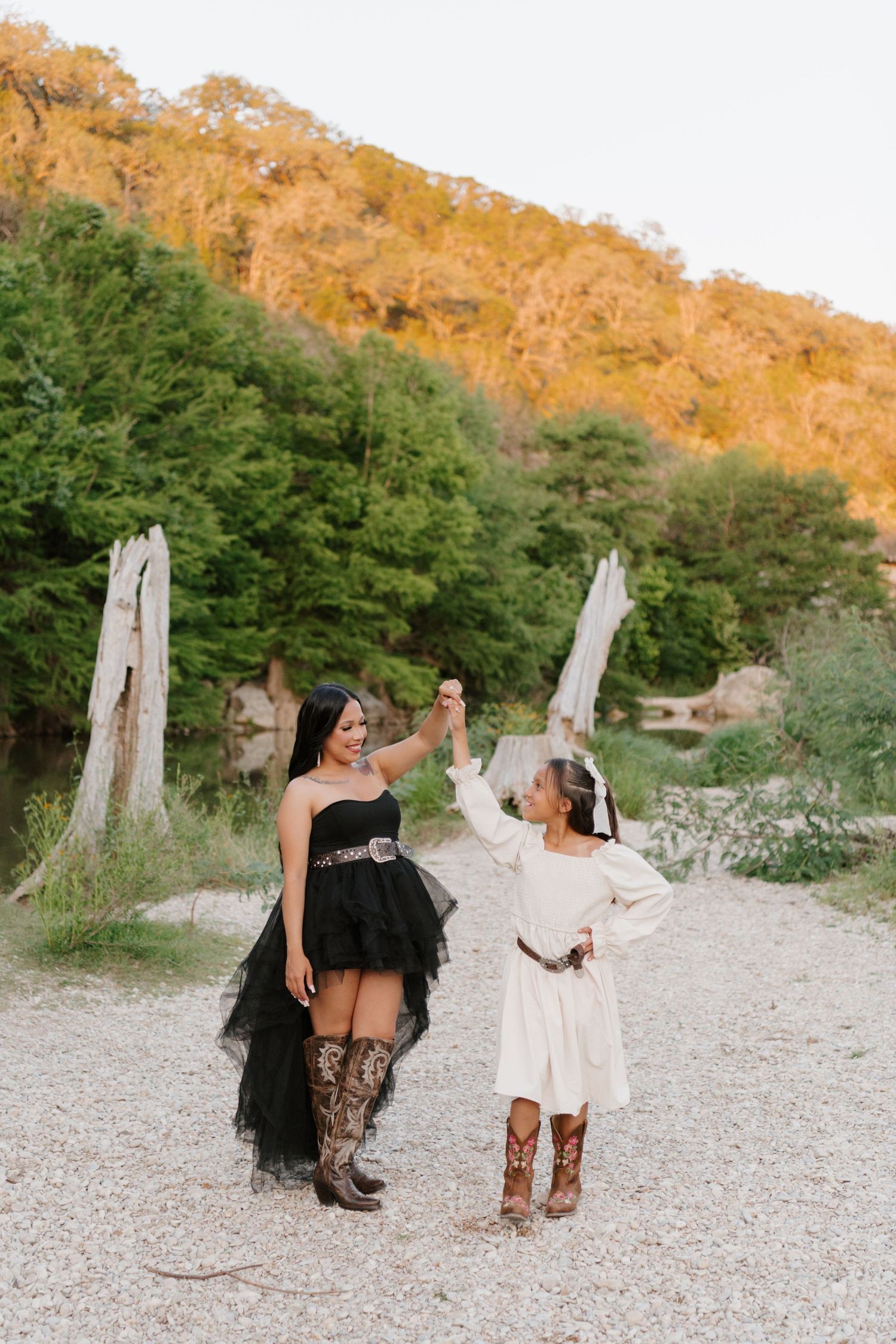 Two people in contrasting black and white outfits with cowboy boots hold hands on a gravel path by sunlit cliffs.