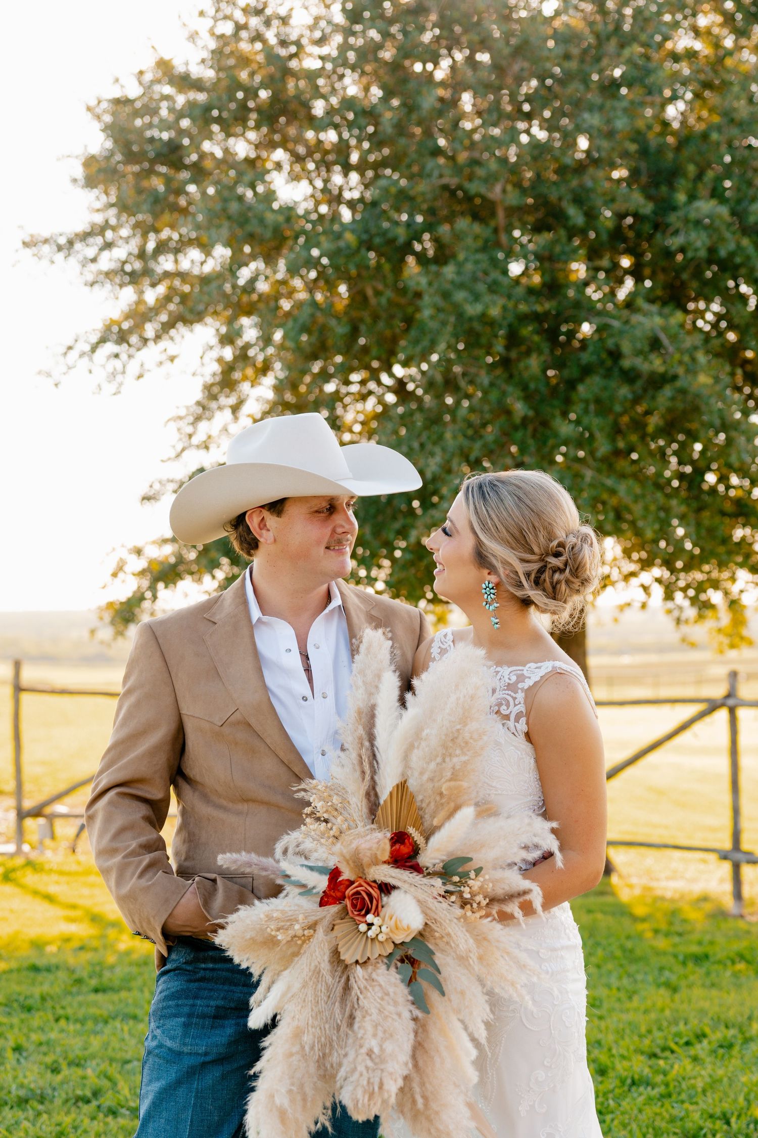 Rustic country wedding couple embracing in sunlit field with pampas grass bouquet.