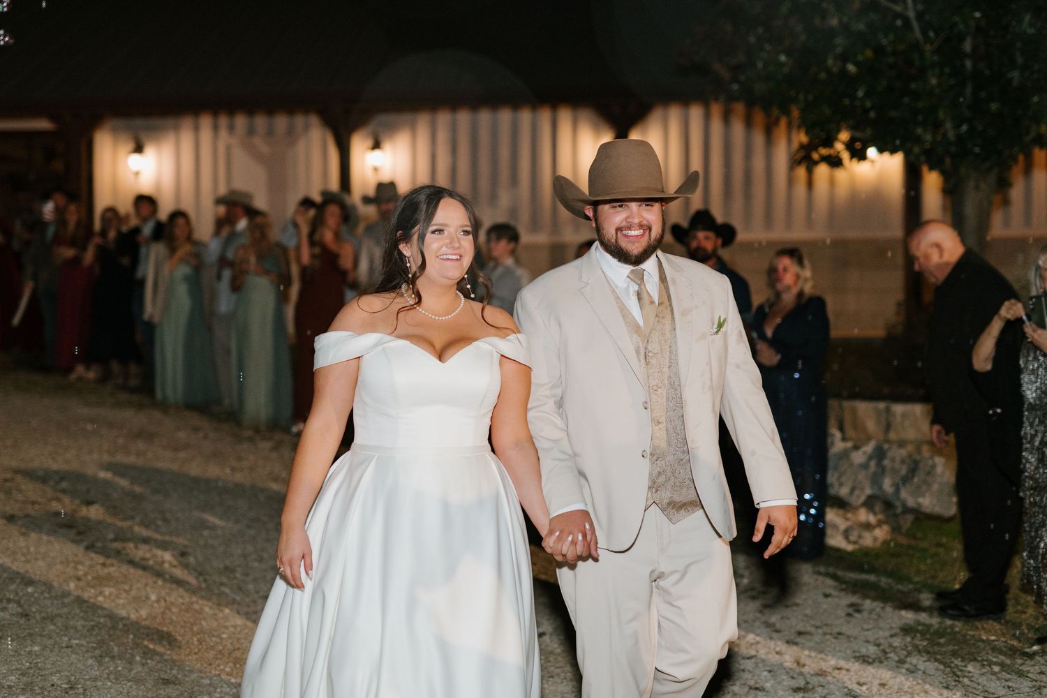 A couple walks hand in hand at their evening wedding celebration, with the bride in white and groom in beige attire.