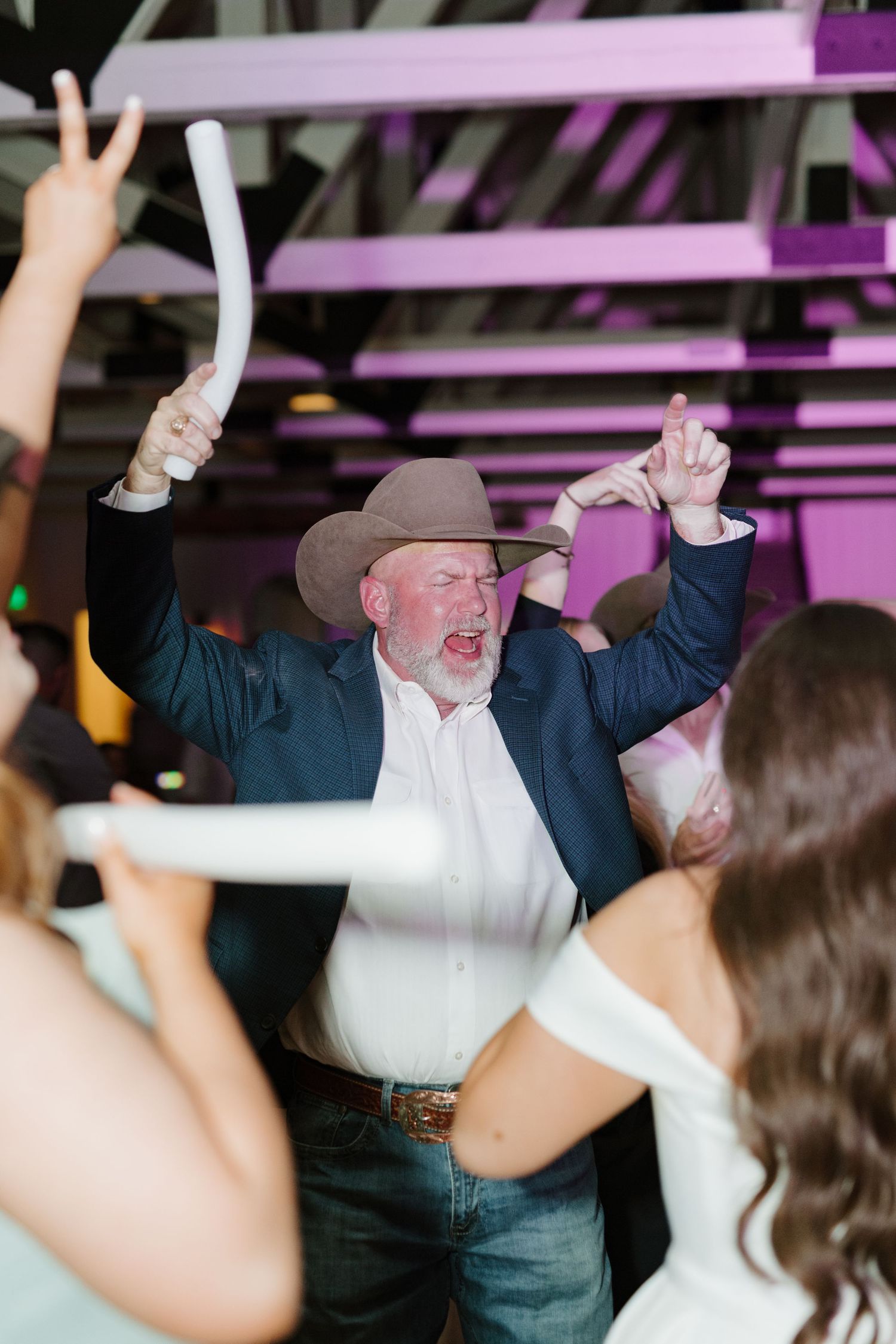 A man in a cowboy hat and jeans dances enthusiastically under purple lighting at what appears to be a celebration.