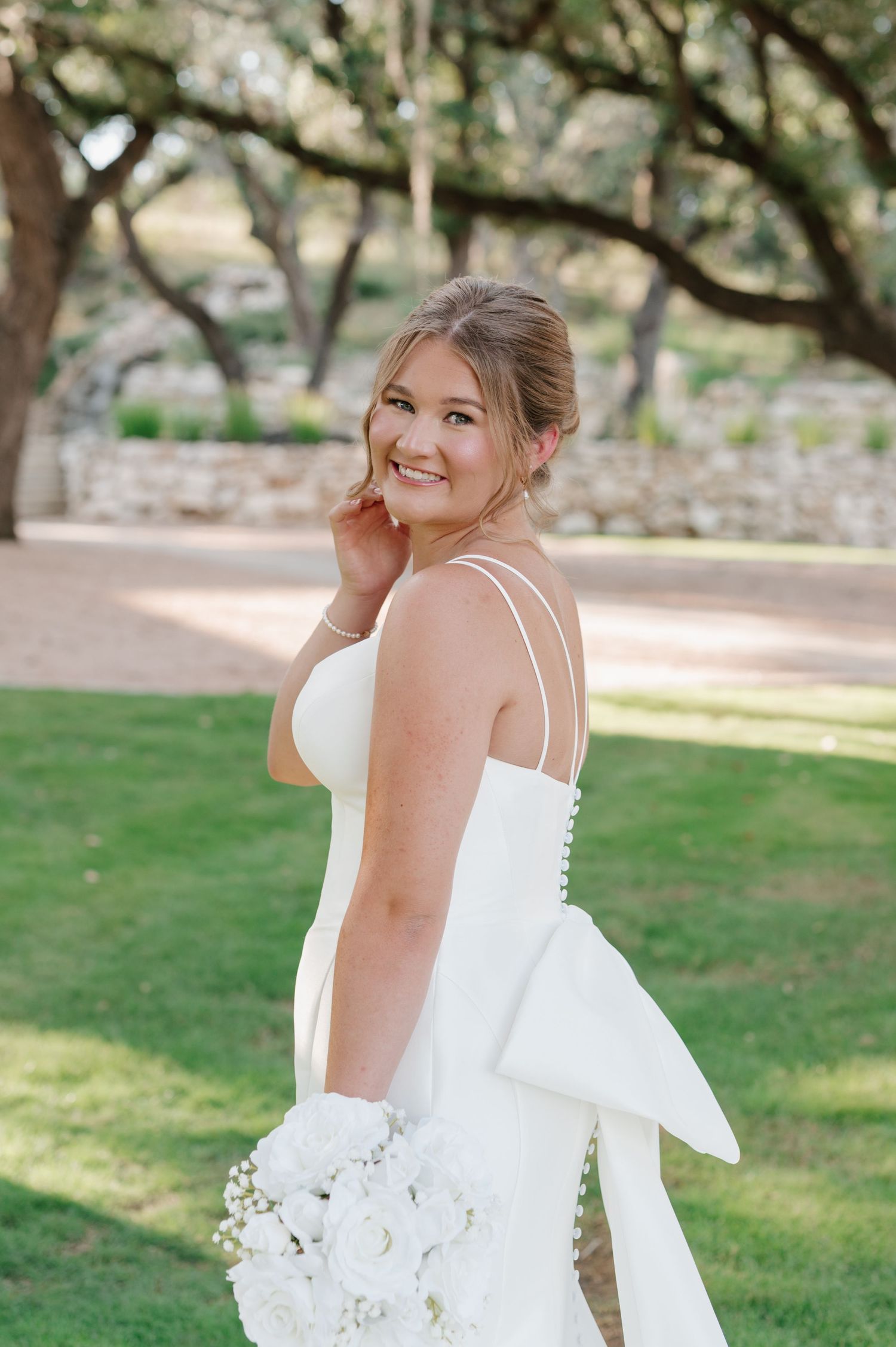 A bride in a sleeveless white gown poses outdoors under oak trees on her wedding day.