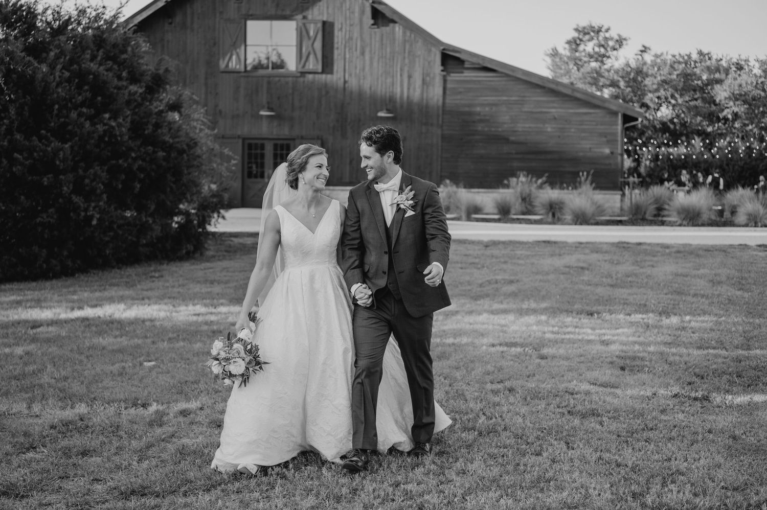 A couple in formal wedding attire walking together on a grassy field at sunset.