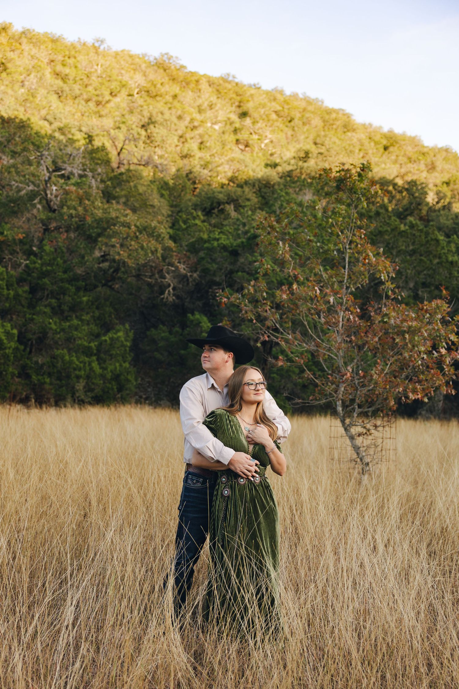 A couple embraces romantically in a golden field of tall grass at sunset with trees in the background.
