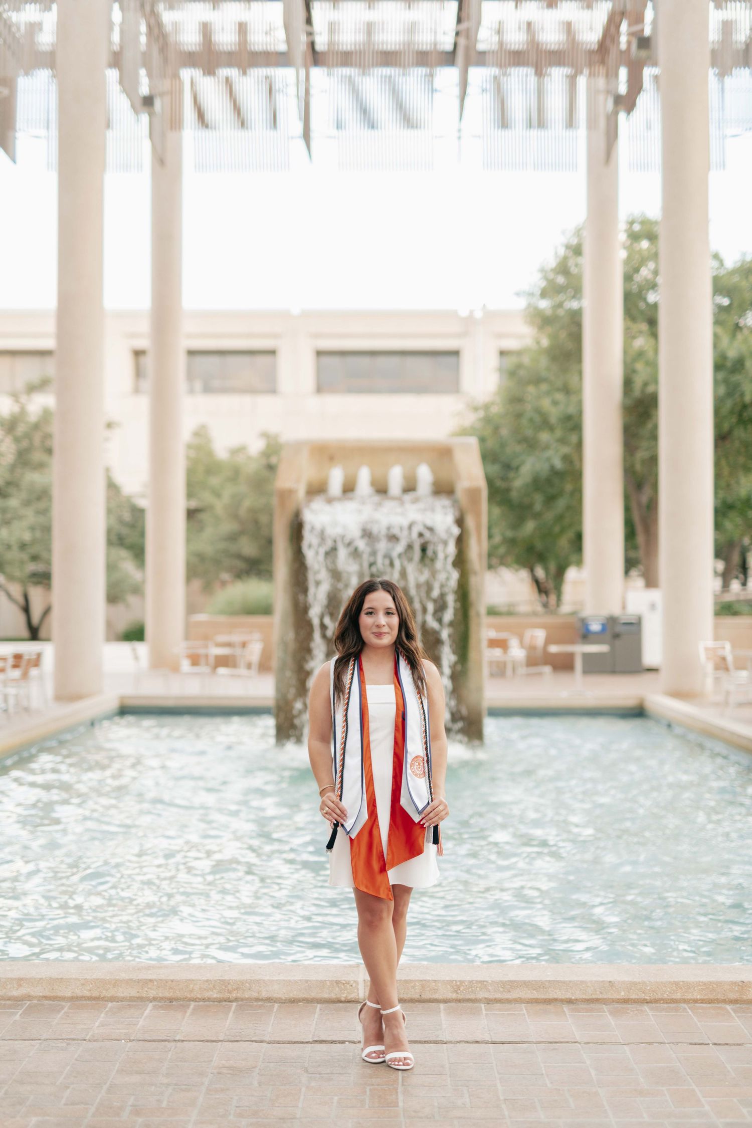 Graduate in white dress and stole stands by decorative fountain.