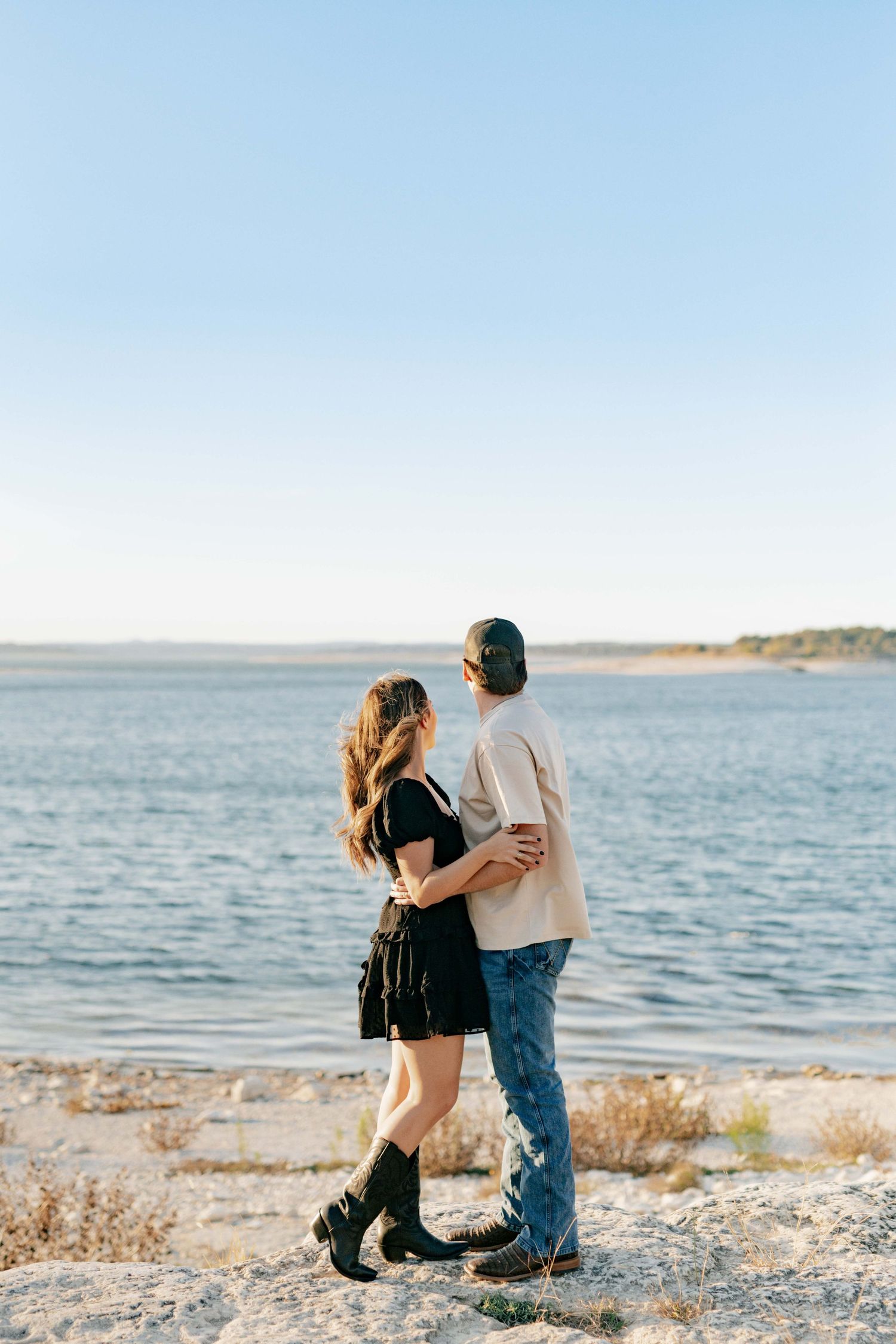 A romantic couple embraces while standing on a rocky shoreline overlooking a serene lake at sunset.