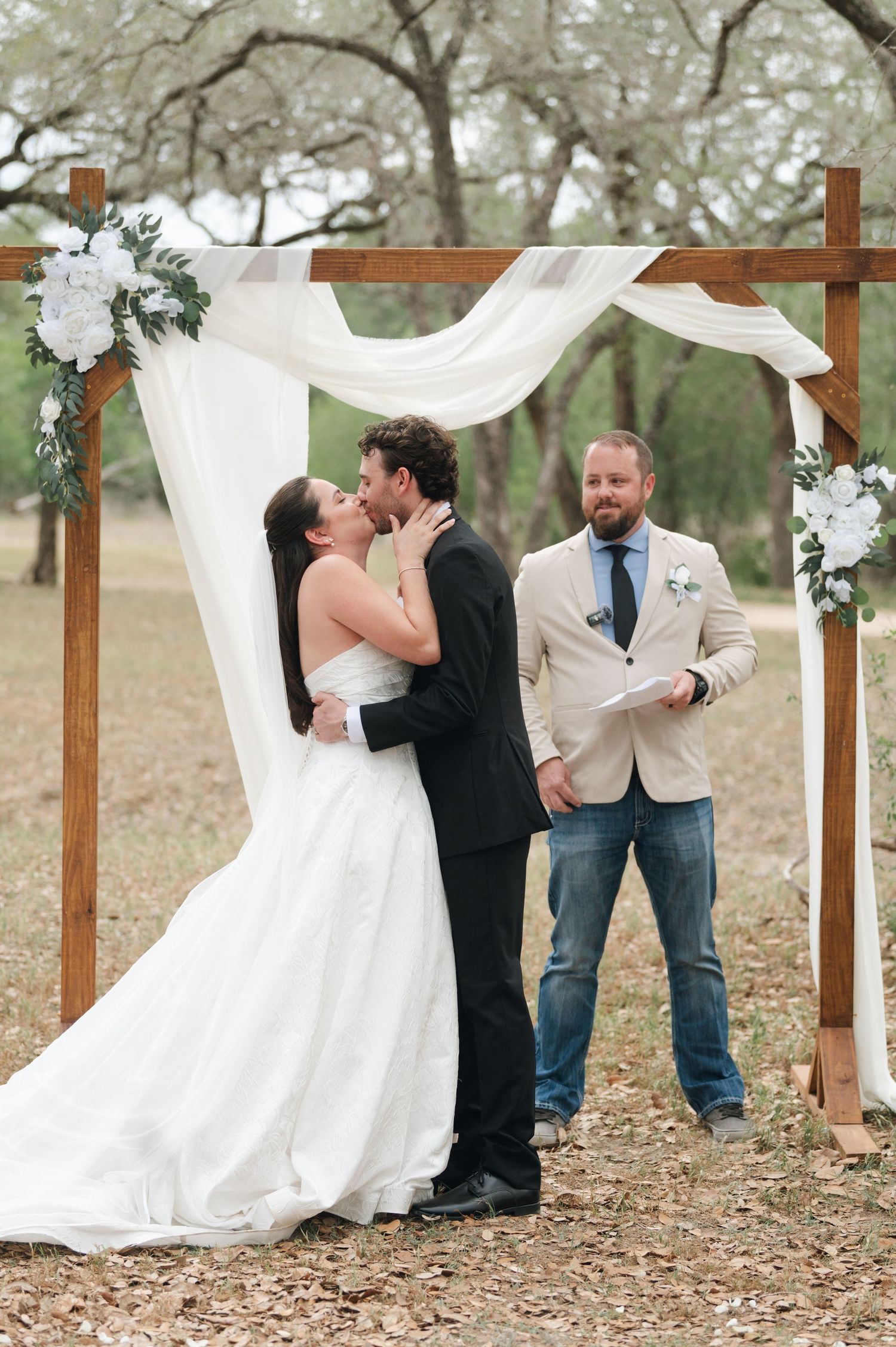 Bride and groom share first kiss under rustic wooden arch with white draping and floral accents at outdoor wedding ceremony.