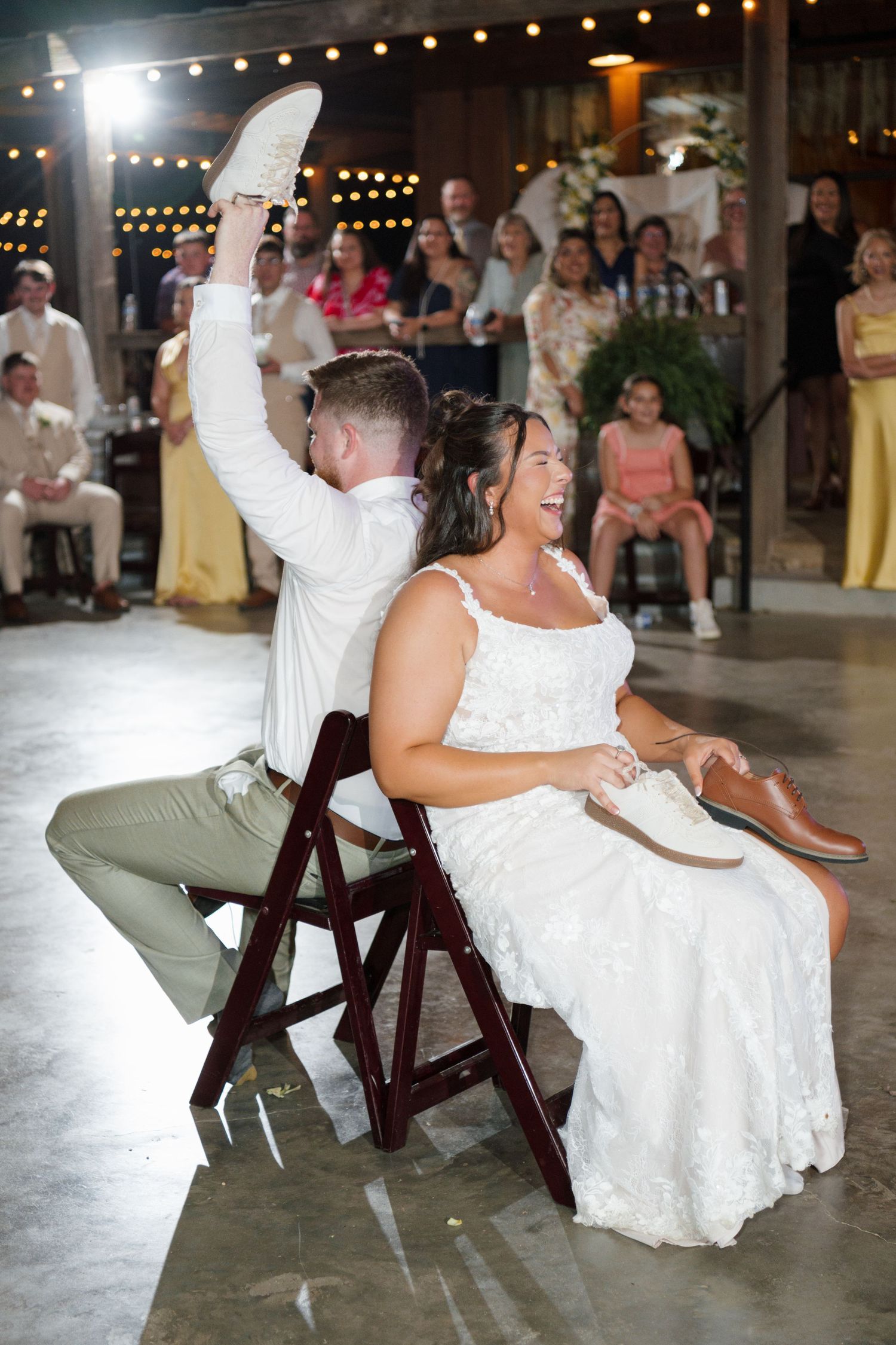 Bride and groom laugh during shoe game at rustic barn wedding reception with guests watching.