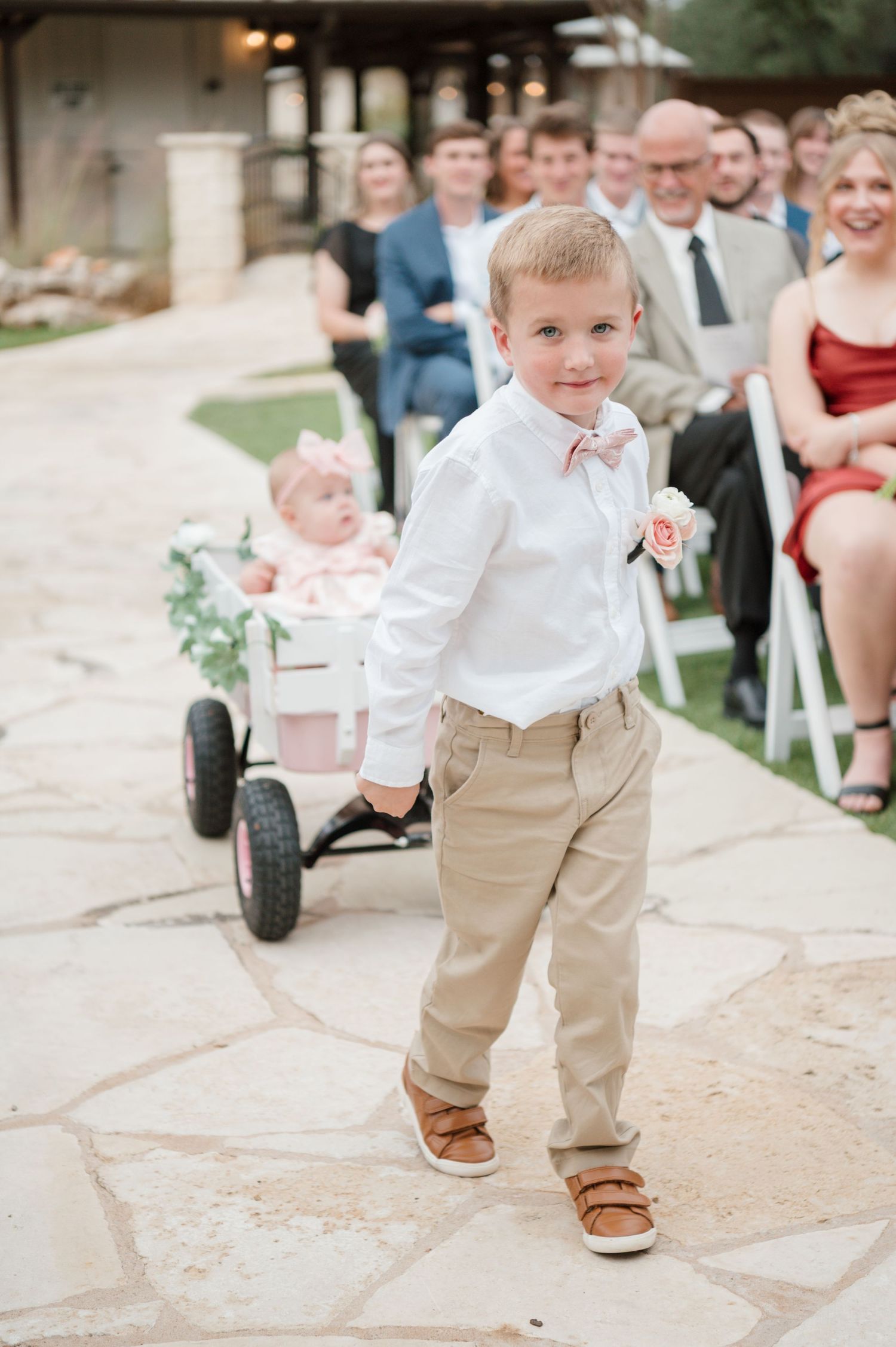 Young ring bearer in white shirt and khakis walks down stone path at outdoor wedding ceremony.